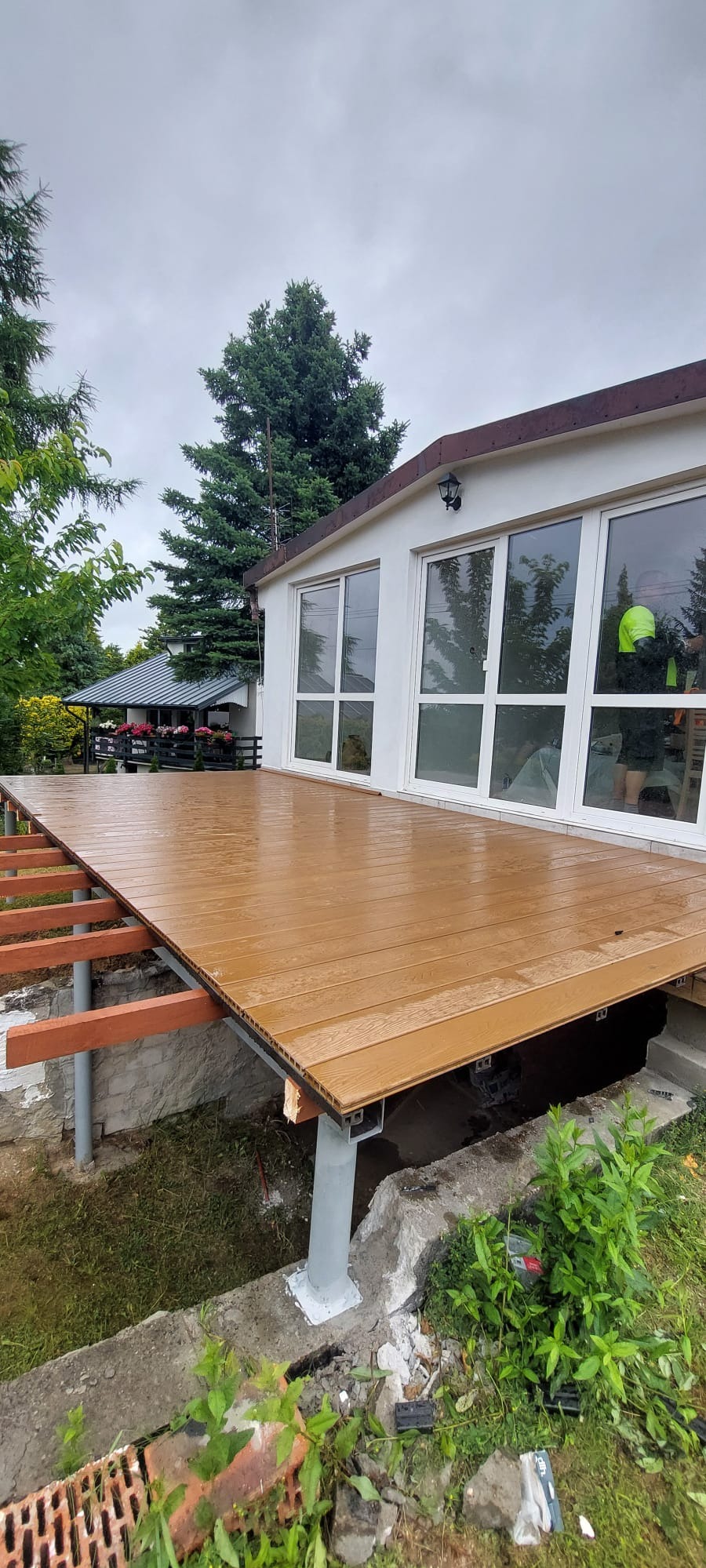 Nowy, brązowy taras kompozytowy przy białym budynku z dużymi oknami, wsparty na metalowych słupach. Widoczne elementy konstrukcyjne pod tarasem.