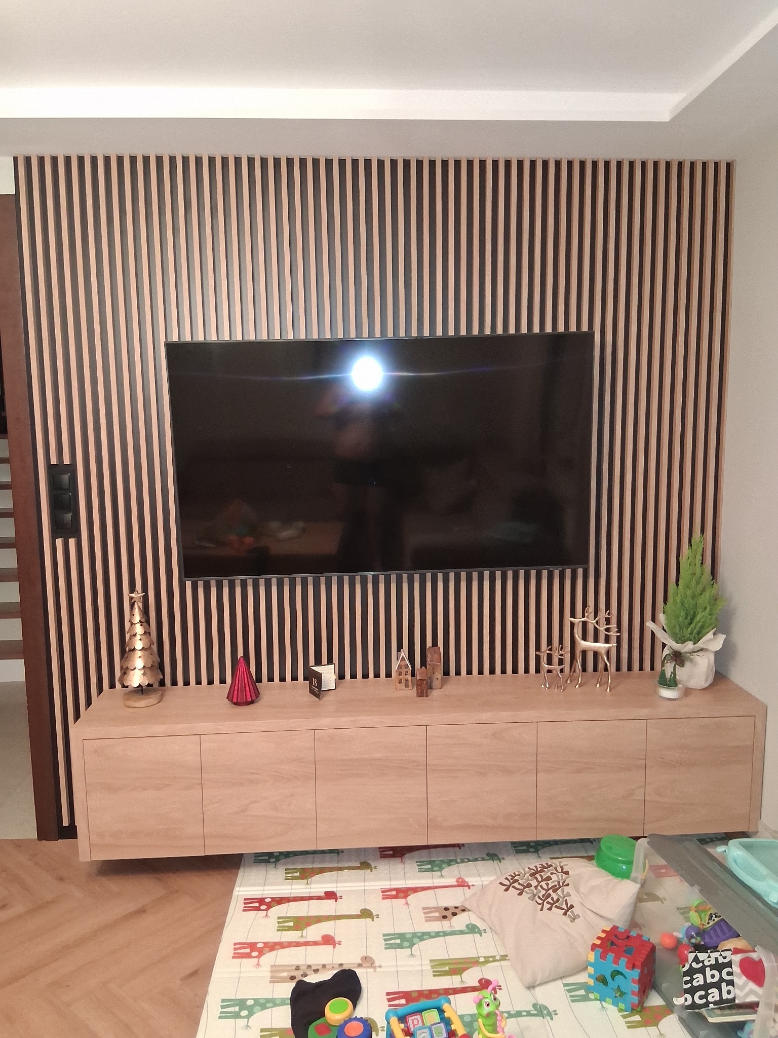 Drewniane lamele na ścianie za telewizorem i szafką RTV w Somoninie. Dekoracje świąteczne na szafce. Jasne drewno, pionowe linie, nowoczesny design.