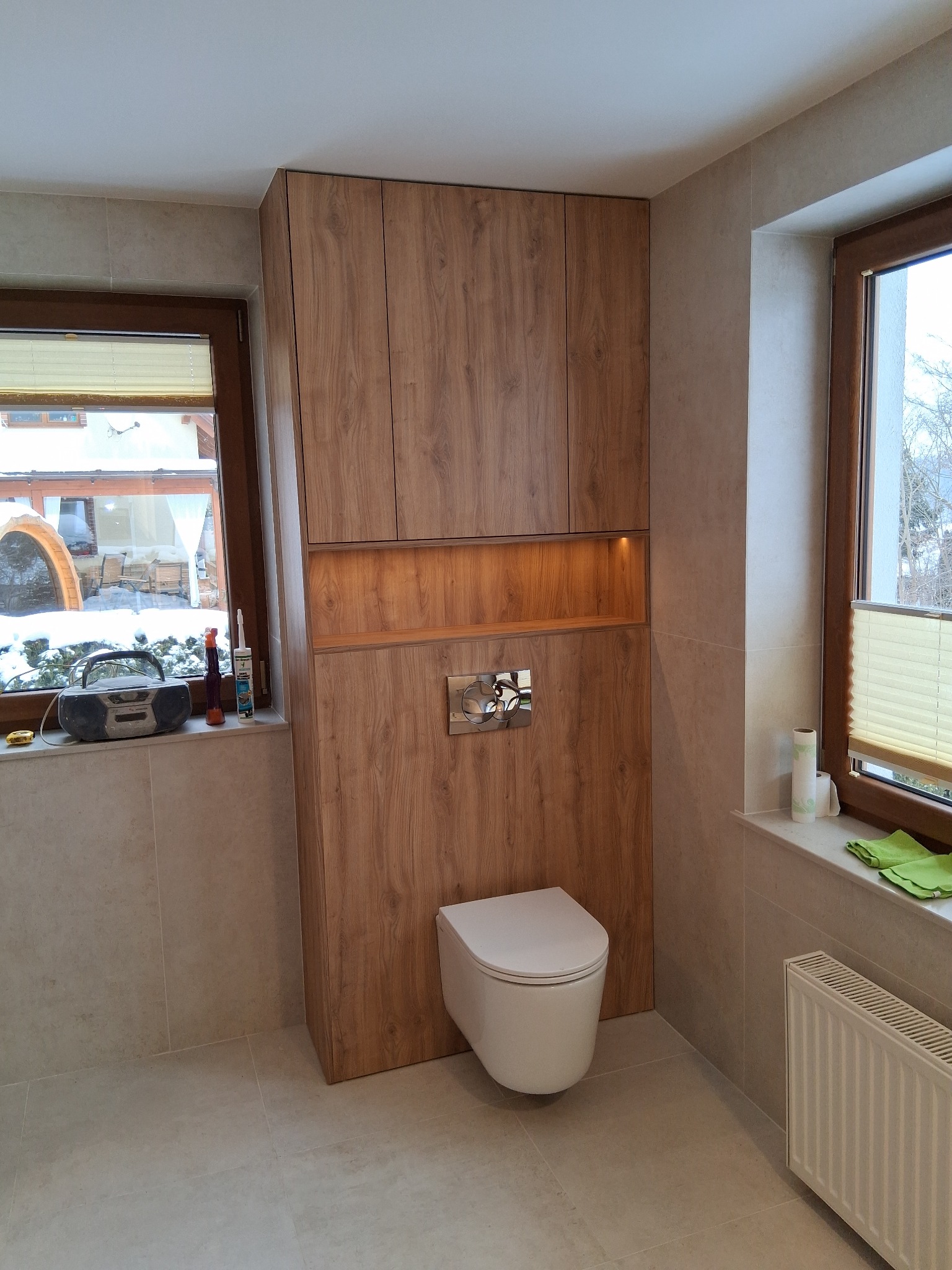 Nowoczesna toaleta z zabudową z drewna w ciepłym odcieniu, z podświetlaną wnęką. Na parapecie radio i akcesoria. Za oknem zimowy krajobraz.