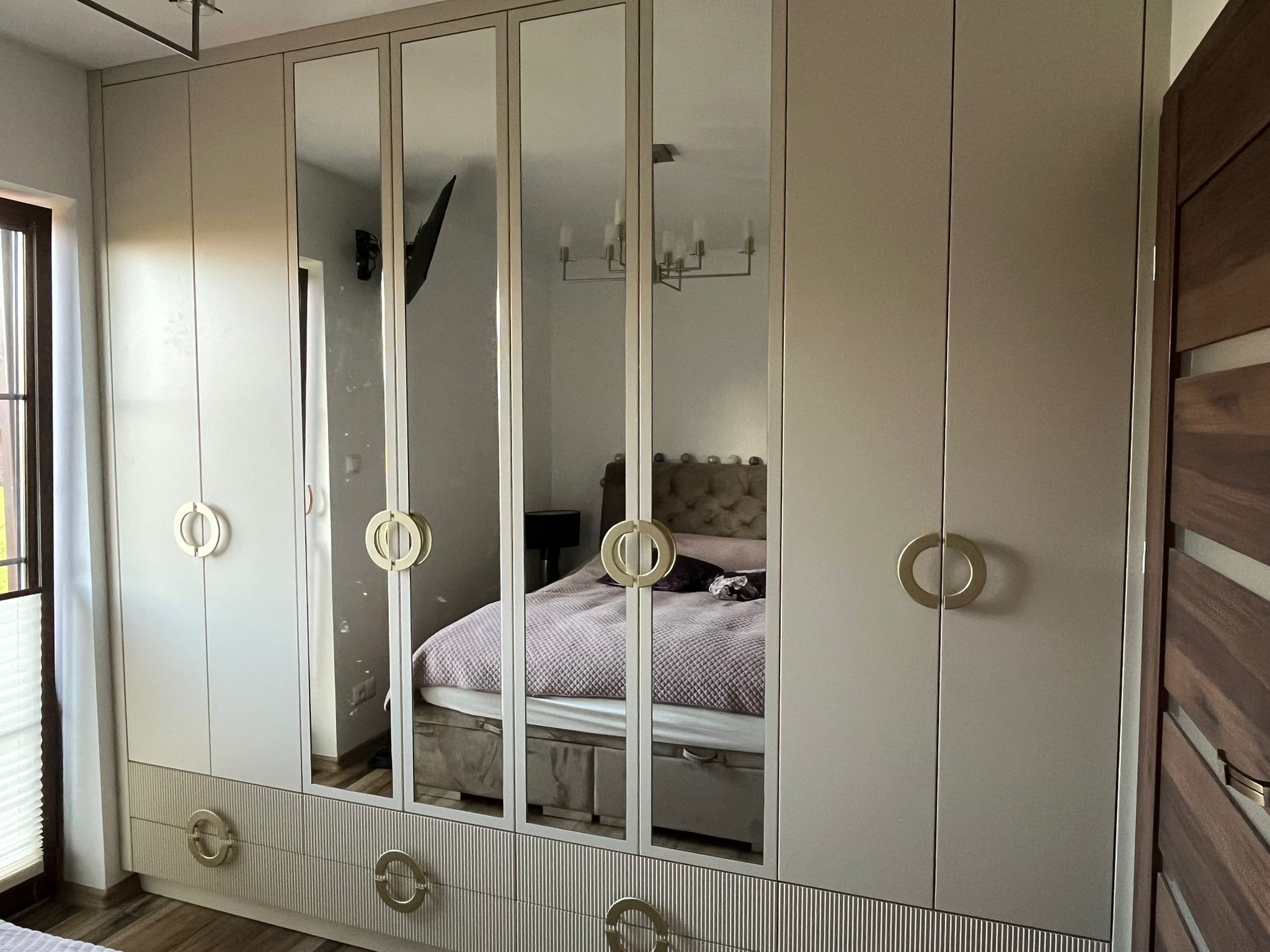 Beżowa szafa z lustrami w sypialni, okrągłe uchwyty, widoczne odbicie łóżka i lampy. Nowoczesny design, pionowe żłobienia na dolnych frontach.