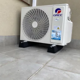 Zewnętrzna jednostka klimatyzacji marki Gree, zamontowana na czarnych podkładkach antywibracyjnych na szarym, wykafelkowanym tarasie przy budynku. Na obudowie widoczna naklejka z pingwinami...