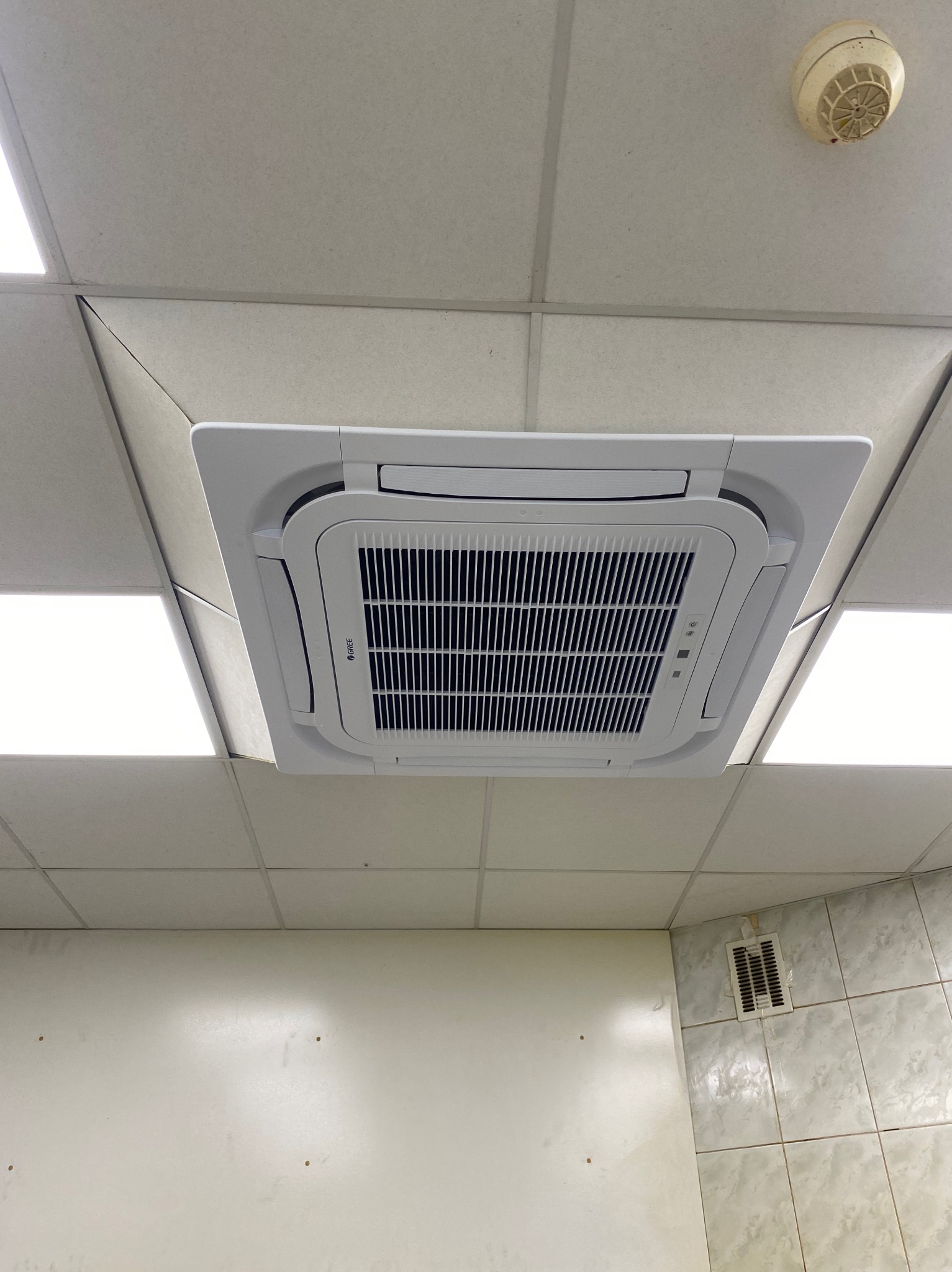 Biała kaseta klimatyzacyjna marki Gree zamontowana w suficie podwieszanym, widok z dołu, obok lampy sufitowe i czujnik dymu.