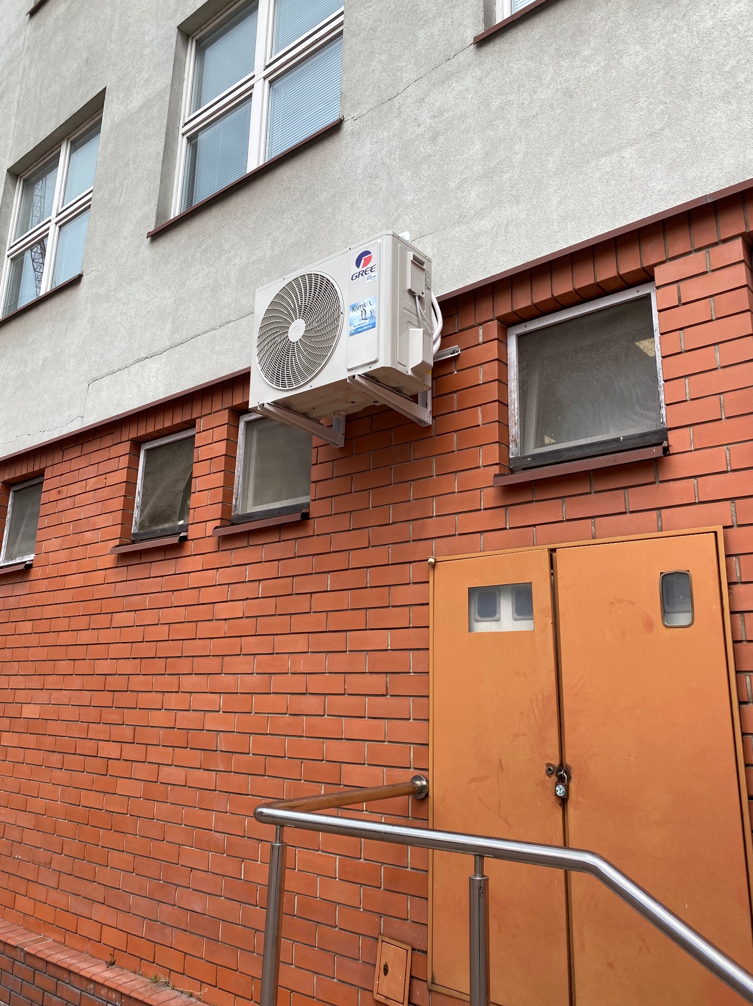 Biały klimatyzator marki Gree zamontowany na ceglanej ścianie budynku z kilkoma oknami i pomarańczowymi drzwiami, widok z dołu.