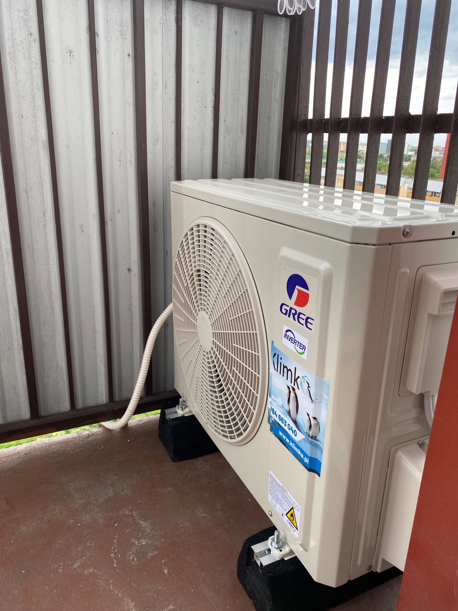 Zewnętrzna jednostka klimatyzacji GREE zamontowana na balkonie, widoczne naklejki firmowe i przewód odprowadzający skropliny.