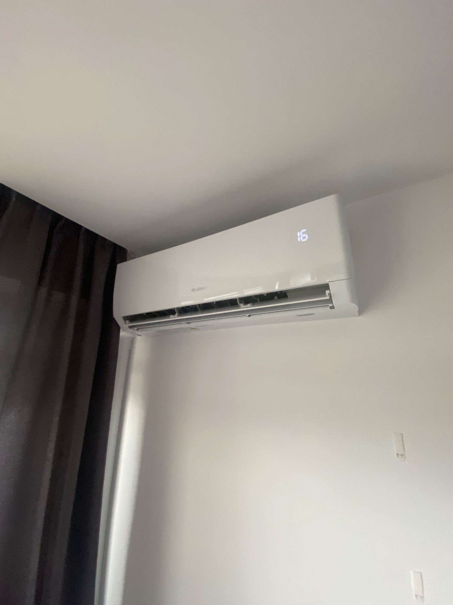 Biały klimatyzator ścienny marki Gree, zamontowany w pomieszczeniu z jasnymi ścianami i ciemnymi zasłonami, wyświetlający temperaturę 16 stopni Celsjusza.