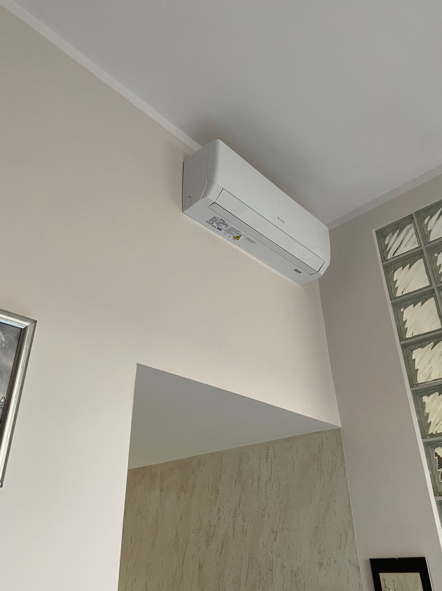 Biały klimatyzator marki Gree zamontowany na ścianie w jasnym pomieszczeniu, widoczne szklane luksfery w ścianie oraz fragment obrazu w ramie.