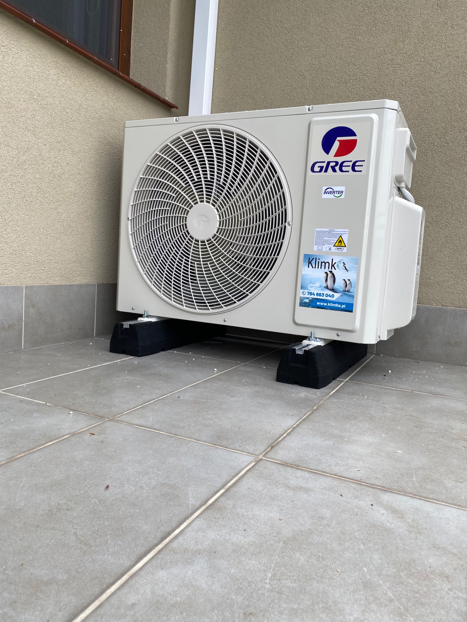 Zewnętrzna jednostka klimatyzacji marki Gree, zamontowana na czarnych podkładkach antywibracyjnych na szarym, wykafelkowanym tarasie przy budynku. Na obudowie widoczna naklejka z pingwinami...