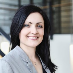 BIELEC ART - fotografia - Uśmiechnięta kobieta z ciemnymi włosami i niebieskimi oczami w szarej marynarce, portret biznesowy na tle nowoczesnych schodów i lamp.