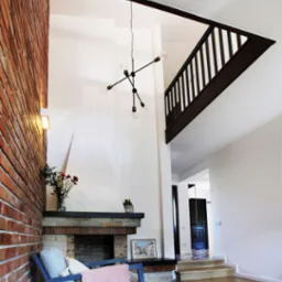 Salon z ceglaną ścianą, kominkiem, nowoczesnym żyrandolem i drewnianą podłogą w jodełkę, widok na antresolę z drewnianą balustradą.