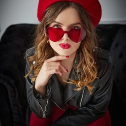 BIELEC ART - fotografia - Kobieta w czerwonym berecie i okularach w kształcie serc pozuje na czarnej sofie. Ubrana w skórzaną kurtkę, z czerwoną szminką i manicure.