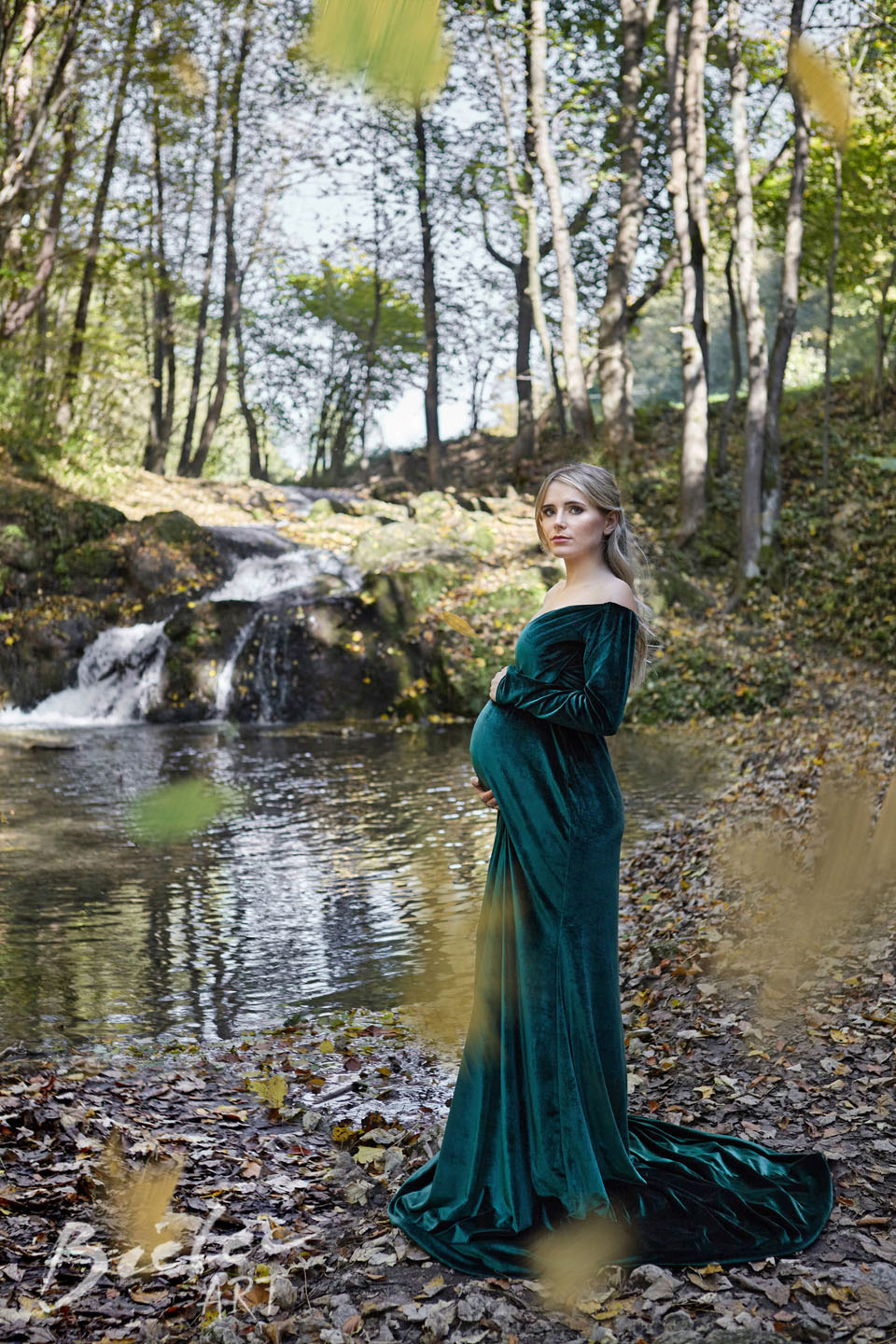 Kobieta w ciąży w szmaragdowej, aksamitnej sukni pozuje w lesie w Krakowie, obok strumienia z małym wodospadem, jesienne liście na ziemi.