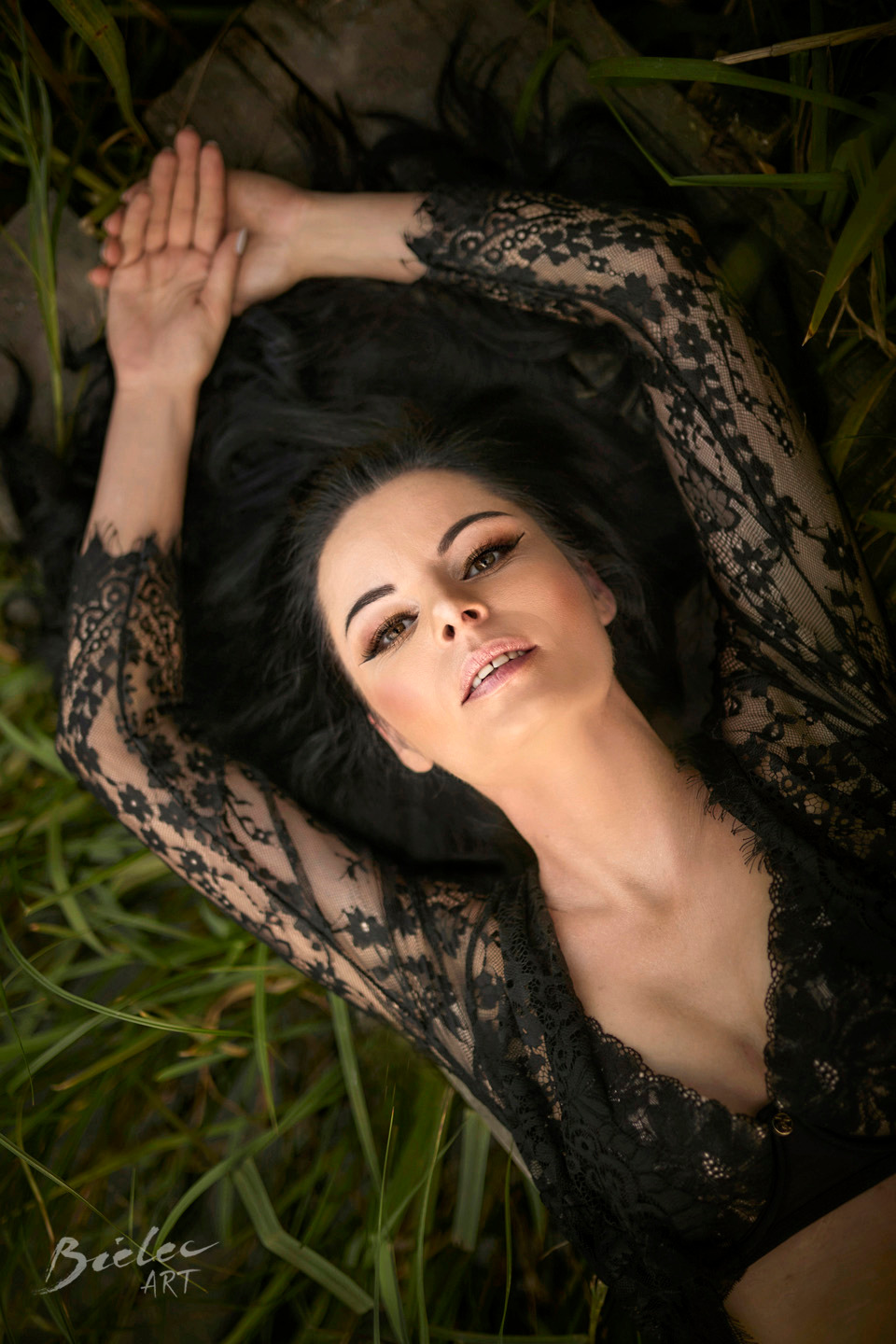 Portret kobiety w czarnej koronkowej sukience leżącej na trawie, z rękami uniesionymi nad głową, skierowana twarzą do góry, z delikatnym makijażem i rozpuszczonymi, ciemnymi włosami.