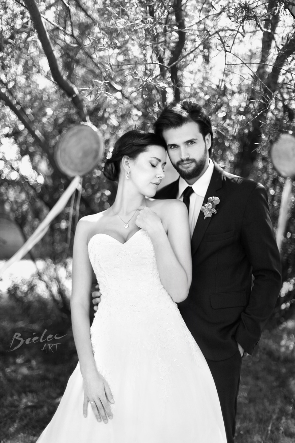 Czarno-białe zdjęcie pary młodej: panna młoda w sukni bez ramiączek z delikatną biżuterią, pan młody w ciemnym garniturze z butonierką, pozujący na tle drzew i dekoracyjnych balonów.