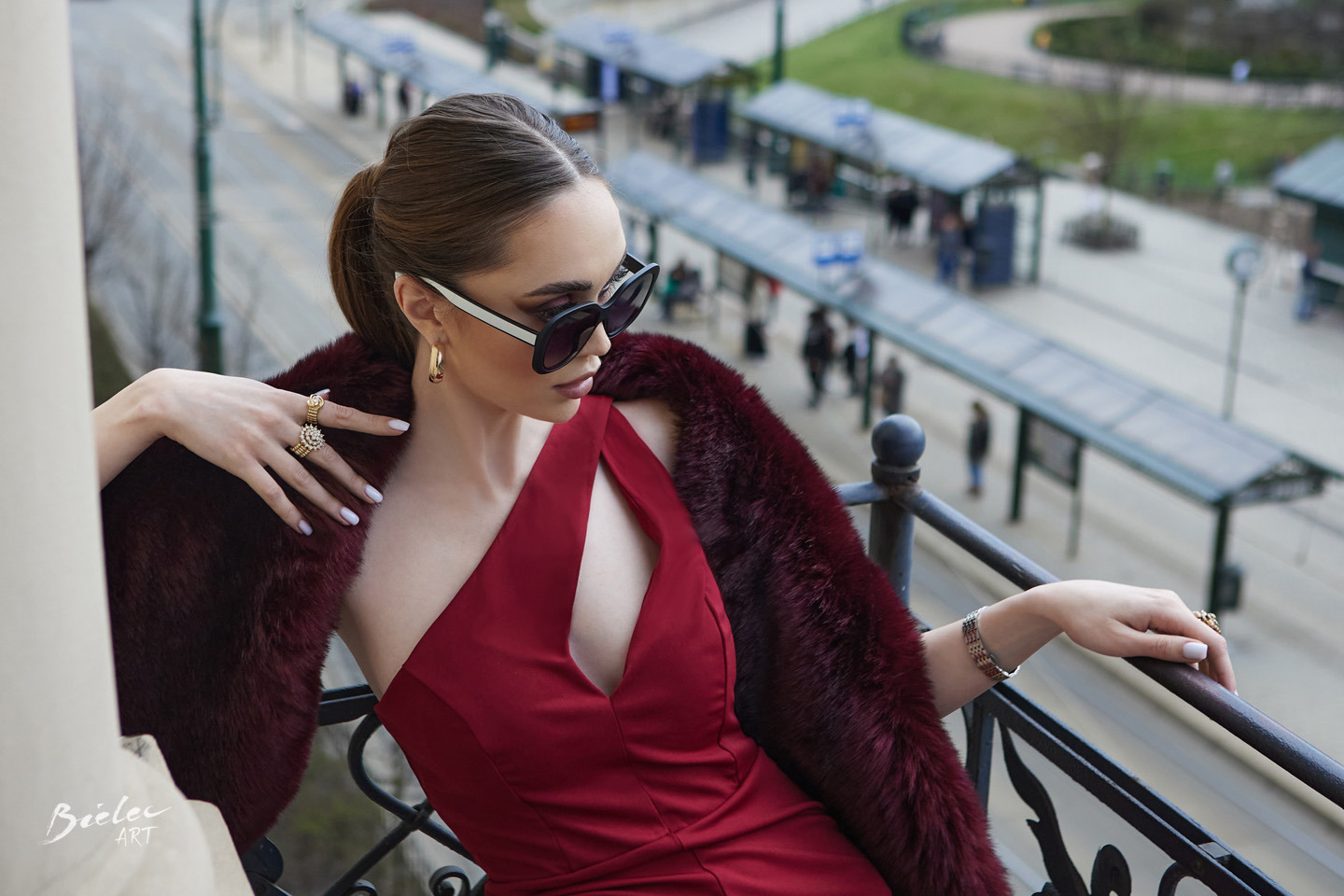 Kobieta w czerwonej sukience i futrzanym szalu, pozująca na balkonie z widokiem na przystanek tramwajowy, z wyraźnie widocznymi złotymi pierścionkami i okularami przeciwsłonecznymi.