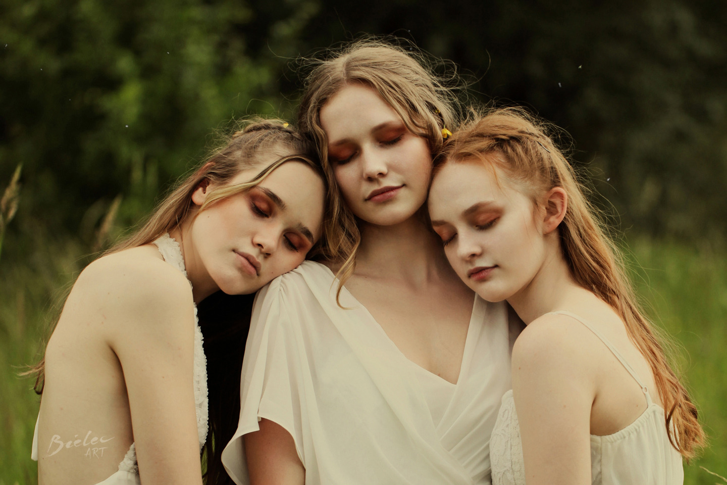 Trzy młode kobiety w zwiewnych białych sukienkach, z zamkniętymi oczami, oparte o siebie w plenerze, subtelny makijaż w odcieniach brązu.