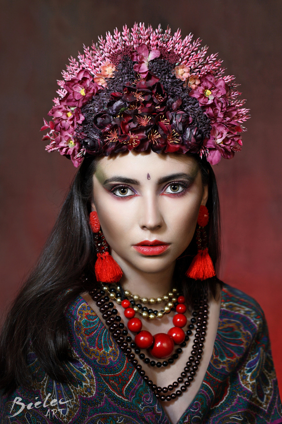 Portret kobiety z ciemnymi włosami, w bogato zdobionym kwiatowym wianku na głowie, czerwonych kolczykach z frędzlami i kilku warstwach naszyjników z koralików, na rozmytym czerwono-brązowym tle.