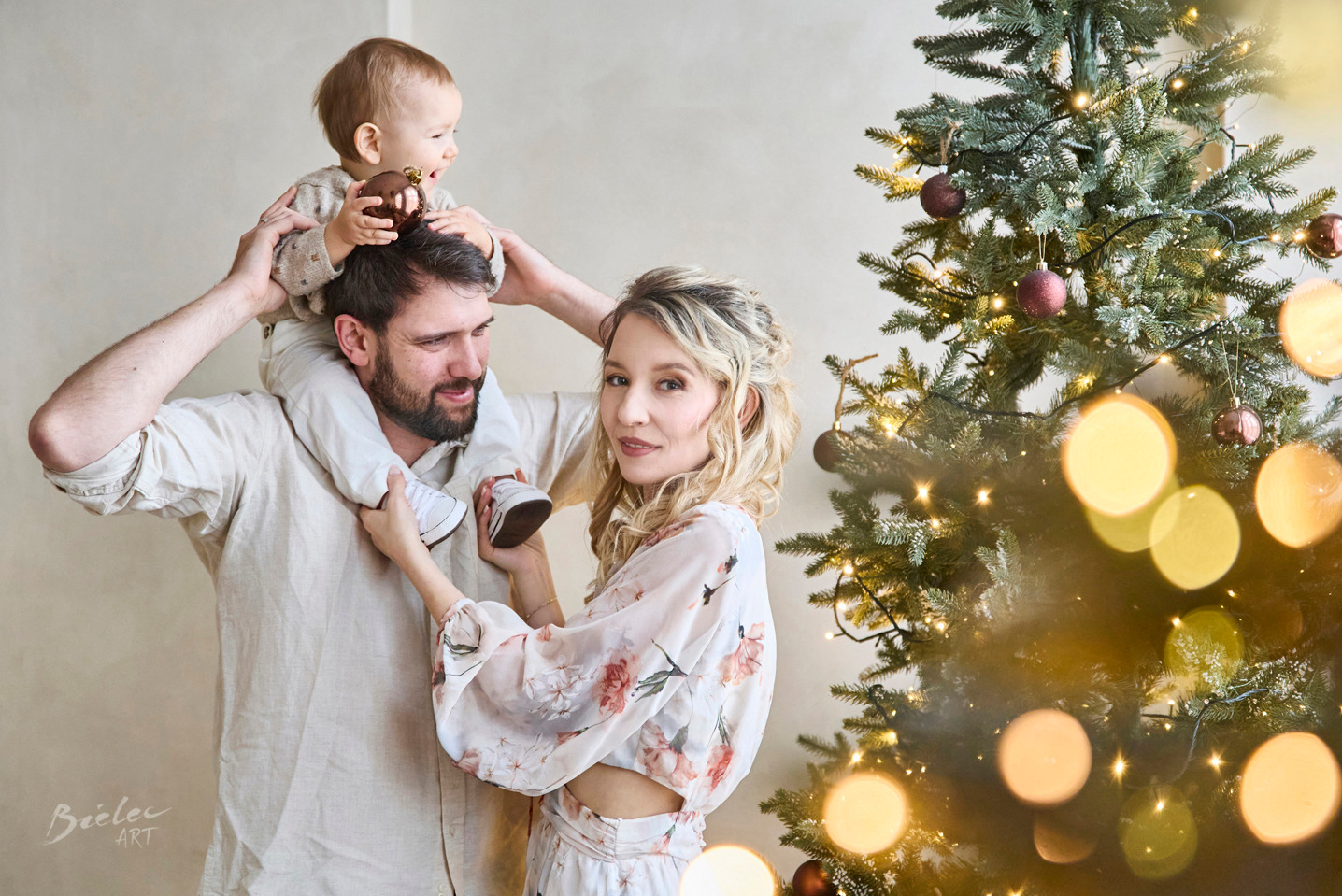 Kraków: Rodzinna sesja świąteczna z tatą trzymającym dziecko na ramionach, mama trzyma małe buciki, w tle choinka z bombkami, ciepłe oświetlenie.