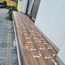 Dominik Trzeciakowski - Układanie płytek imitujących drewno na balkonie z użyciem poziomujących klipsów i klinów; widoczna elewacja budynku i kostka brukowa.