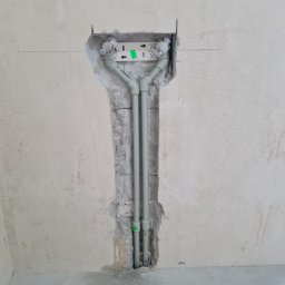 Dominik Trzeciakowski - Instalacja wodna z rur PP-R w bruździe ściany, widoczne złącza i uchwyty montażowe, ściana w stanie surowym przed wykończeniem, podłoga betonowa.