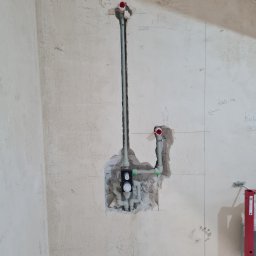 Dominik Trzeciakowski - Surowa ściana z instalacją wodną w trakcie montażu, widoczne rury PCV, oznaczenia wymiarów naniesione ołówkiem na ścianie, oraz fragment poziomicy.