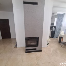 Dominik Trzeciakowski - Nowoczesny salon z kominkiem obudowanym szarą strukturą, jasna podłoga z płytek, widoczny fragment jadalni z niebieskimi zasłonami.