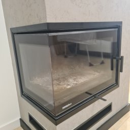 Dominik Trzeciakowski - Zamknięty, narożny kominek z czarną ramą i szklanymi drzwiczkami, widoczny popiół wewnątrz paleniska, ściana wykończona szarym tynkiem strukturalnym, na podłodze jasne płytki.