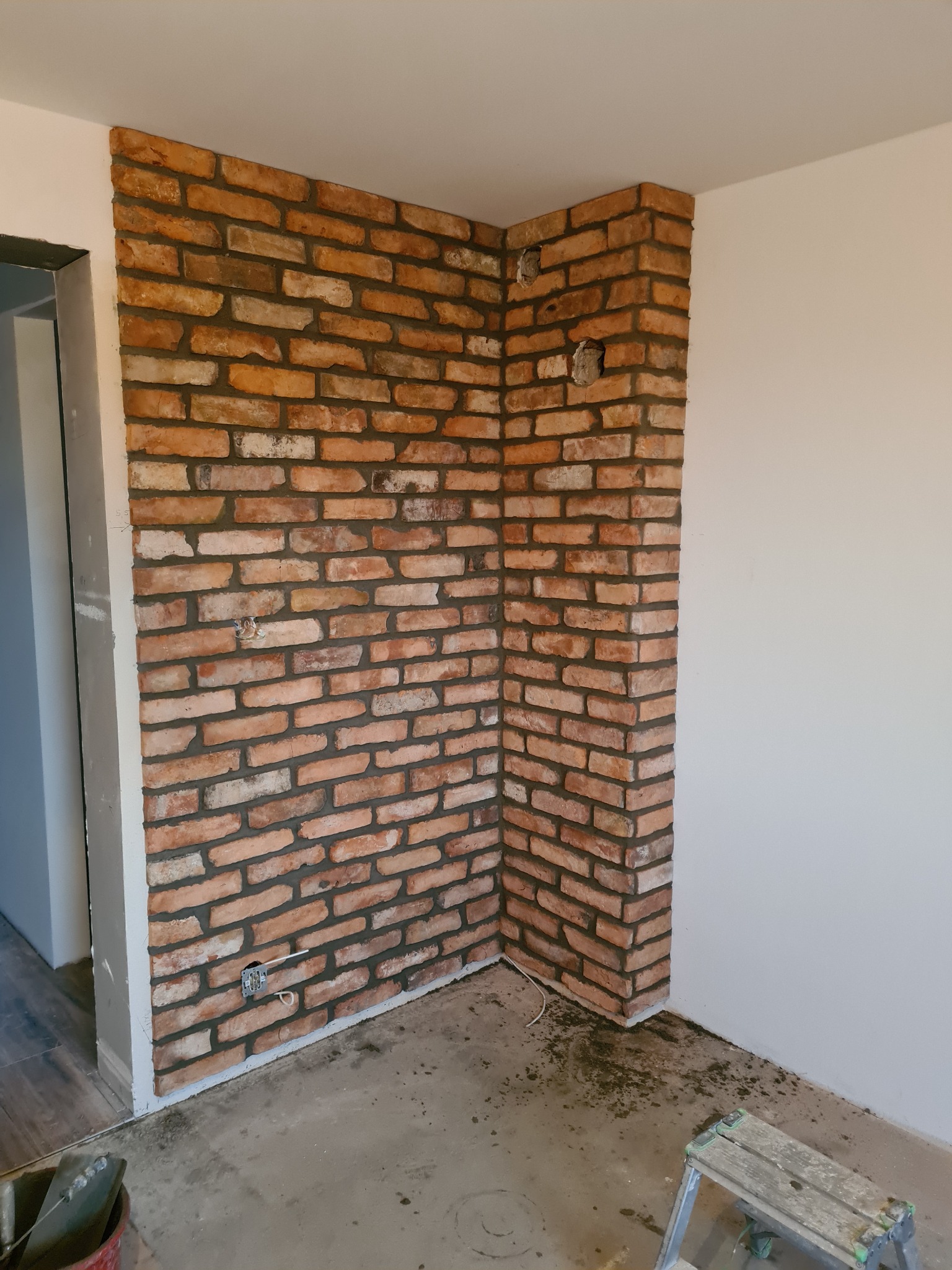 Fragment ściany wykończonej czerwoną cegłą klinkierową w narożniku pomieszczenia, widoczne gniazdko elektryczne, nieukończona podłoga i drabinka malarska.