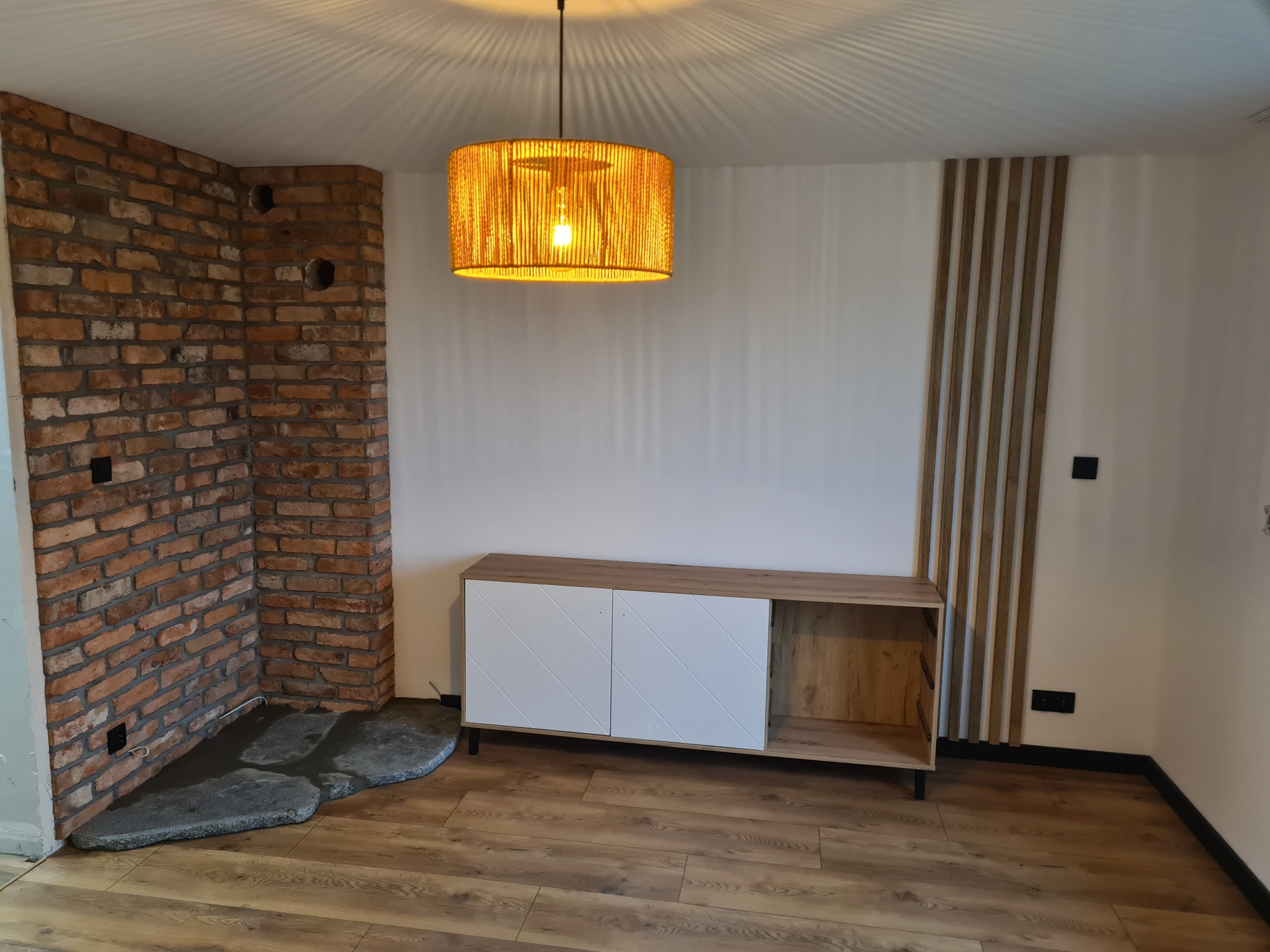 Salon po metamorfozie z ceglaną ścianą, drewnianą podłogą, szafką RTV z białymi frontami i dekoracyjnymi panelami ściennymi, oświetlony wiszącą lampą z abażurem z naturalnego włókna.