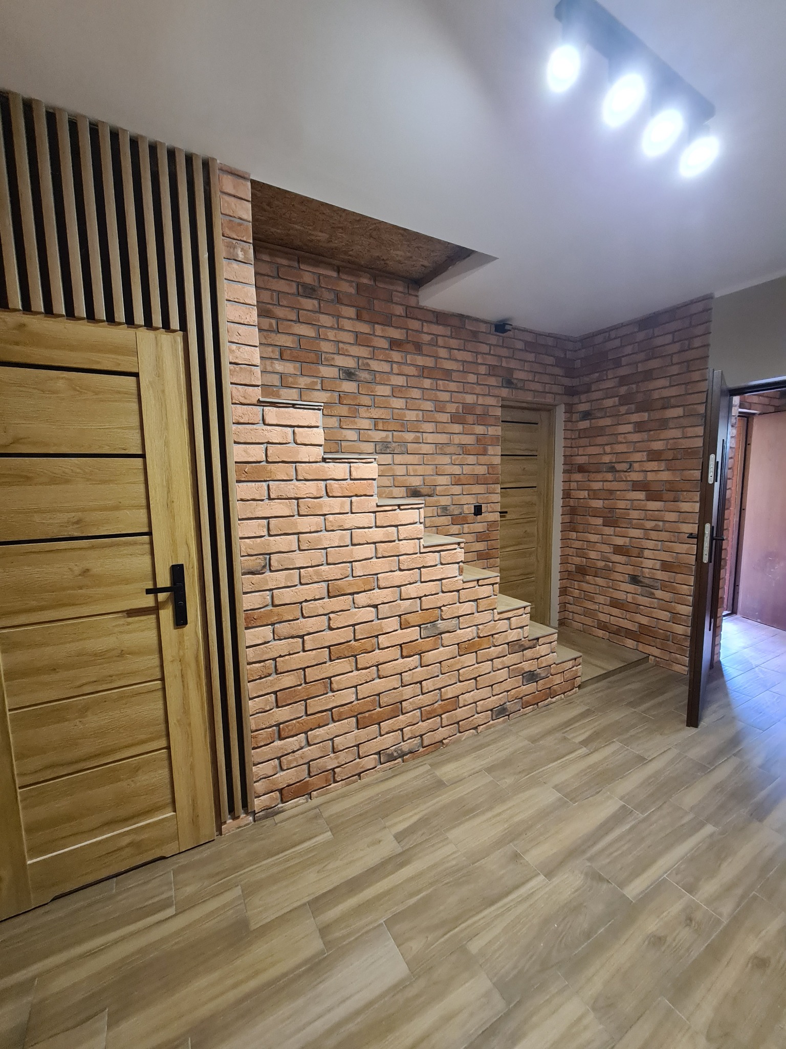 Wnętrze korytarza z ceglaną ścianą, drewnianymi panelami pionowymi i podłogą imitującą drewno, oświetlenie sufitowe punktowe, widoczne schodki z cegieł.