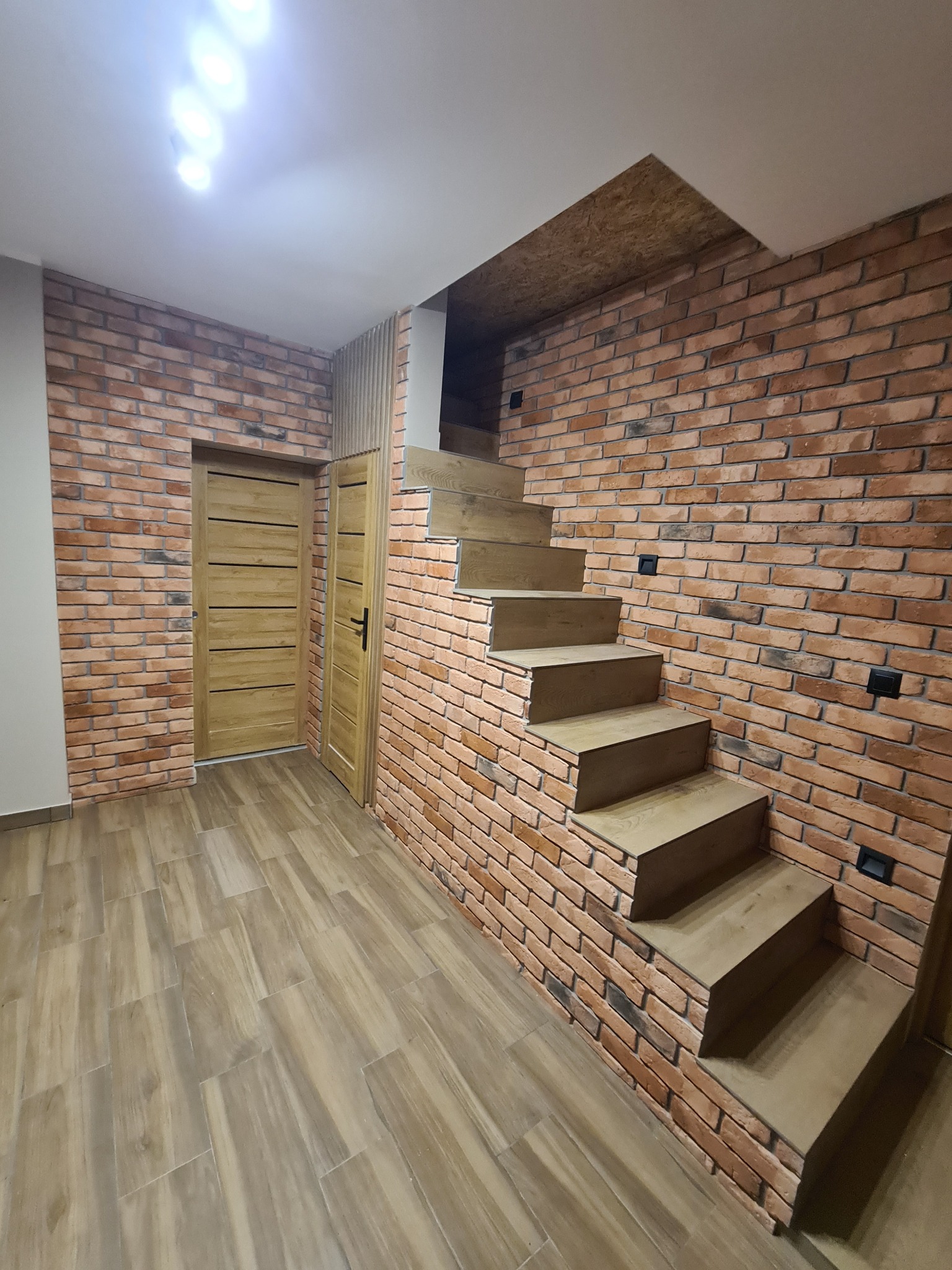 Wykończone wnętrze z ceglaną ścianą, drewnianymi schodami bez poręczy i drewnianymi drzwiami, oświetlone punktowymi lampami sufitowymi, z podłogą imitującą drewno.