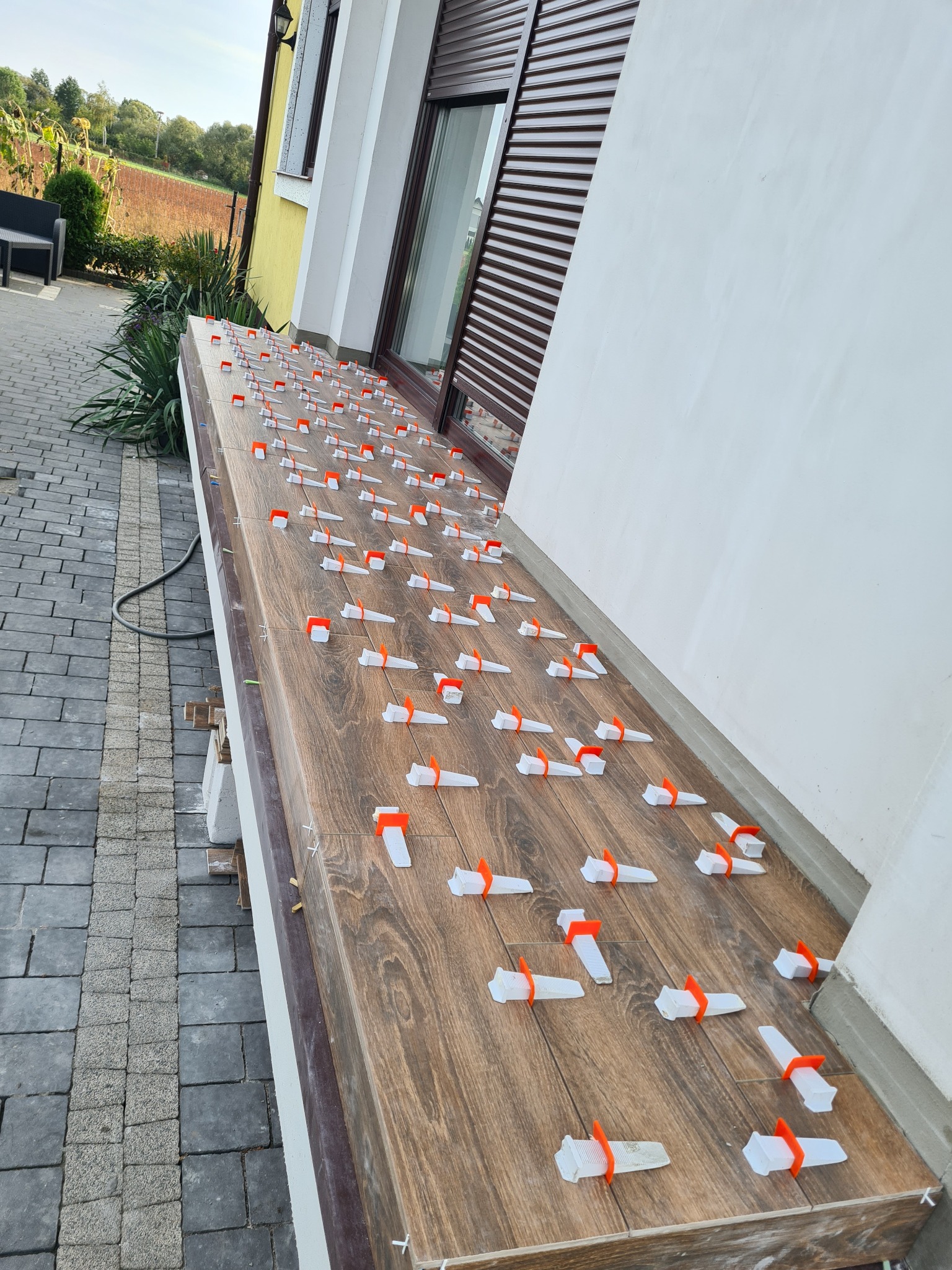 Układanie płytek imitujących drewno na balkonie z użyciem poziomujących klipsów i klinów; widoczna elewacja budynku i kostka brukowa.