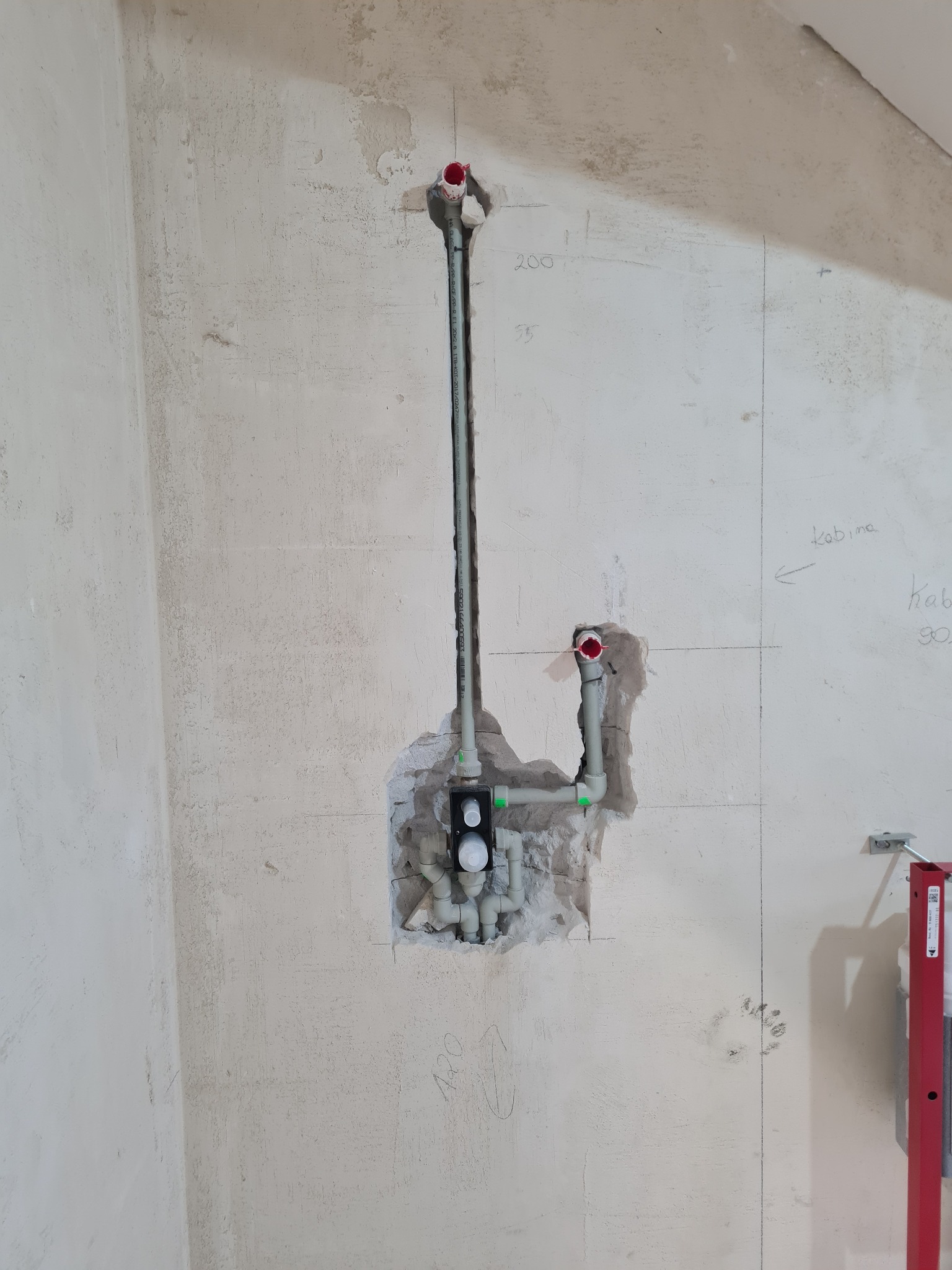 Surowa ściana z instalacją wodną w trakcie montażu, widoczne rury PCV, oznaczenia wymiarów naniesione ołówkiem na ścianie, oraz fragment poziomicy.