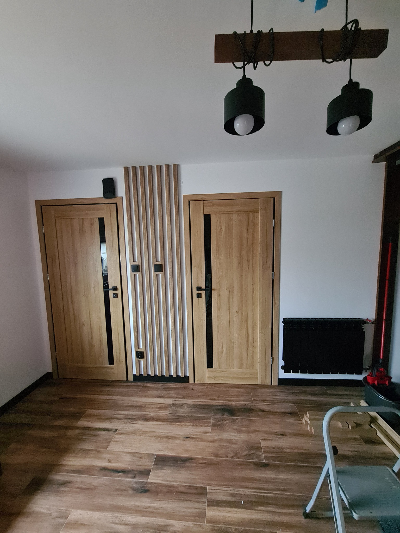 Dwa drewniane panele drzwiowe z czarnymi akcentami, oddzielone pionowymi listwami dekoracyjnymi, na podłodze imitującej drewno, z czarnym grzejnikiem w tle.