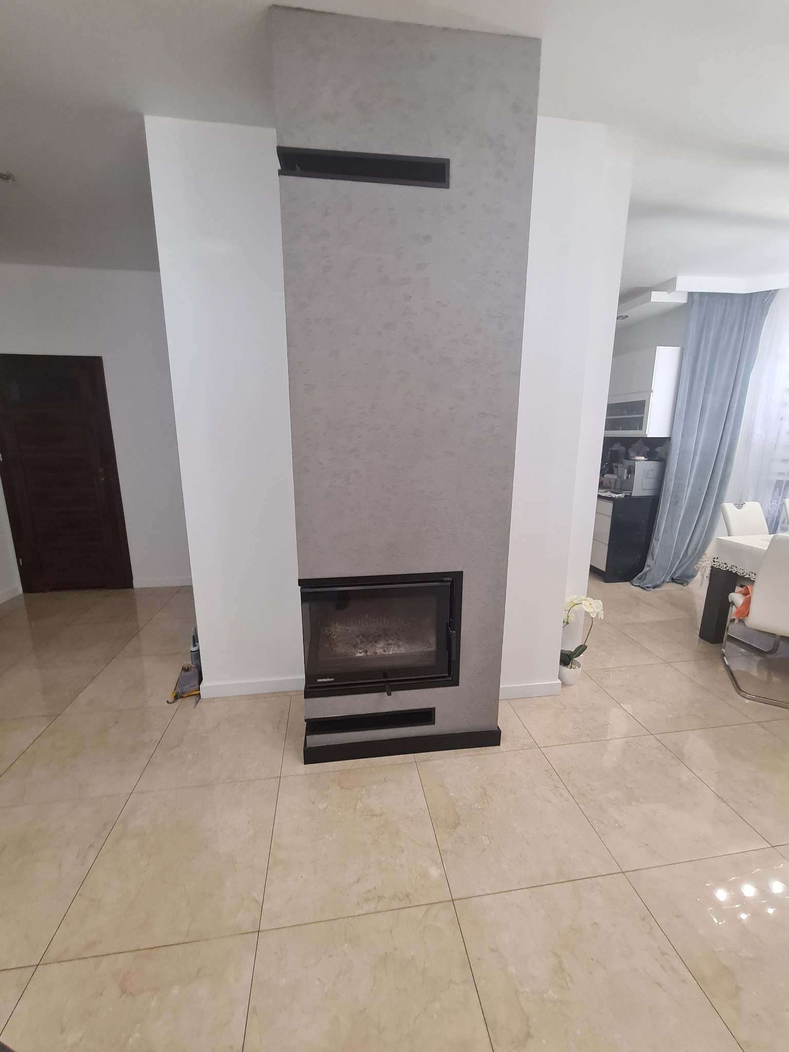 Nowoczesny salon z kominkiem obudowanym szarą strukturą, jasna podłoga z płytek, widoczny fragment jadalni z niebieskimi zasłonami.