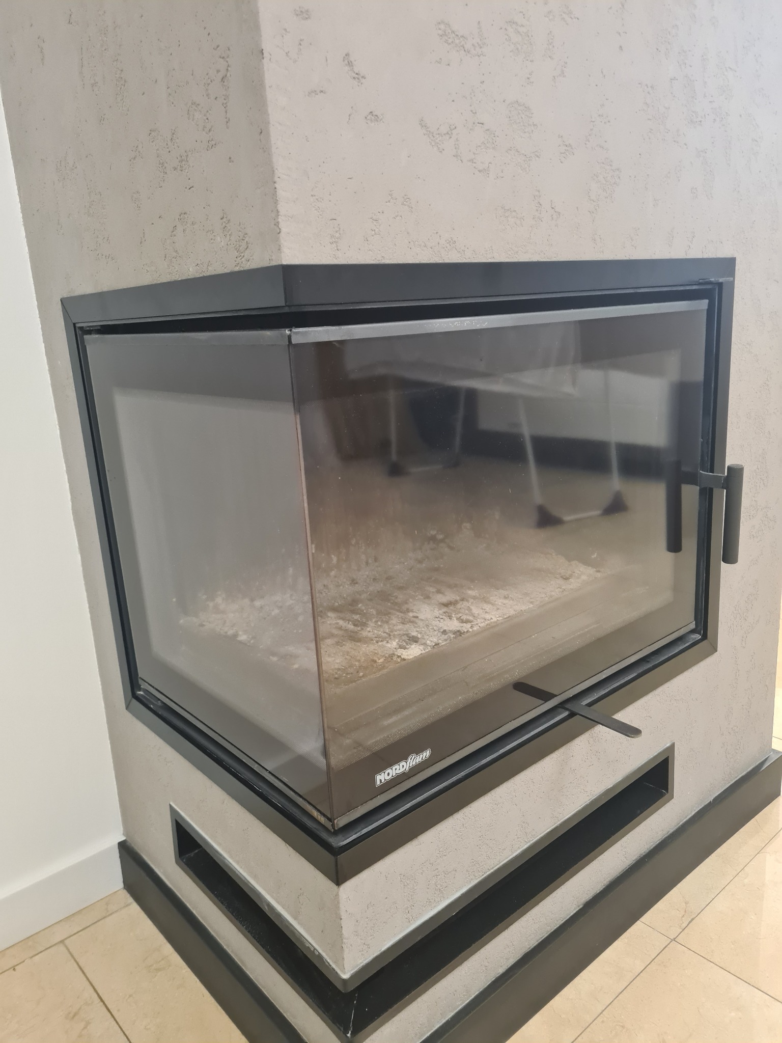 Zamknięty, narożny kominek z czarną ramą i szklanymi drzwiczkami, widoczny popiół wewnątrz paleniska, ściana wykończona szarym tynkiem strukturalnym, na podłodze jasne płytki.