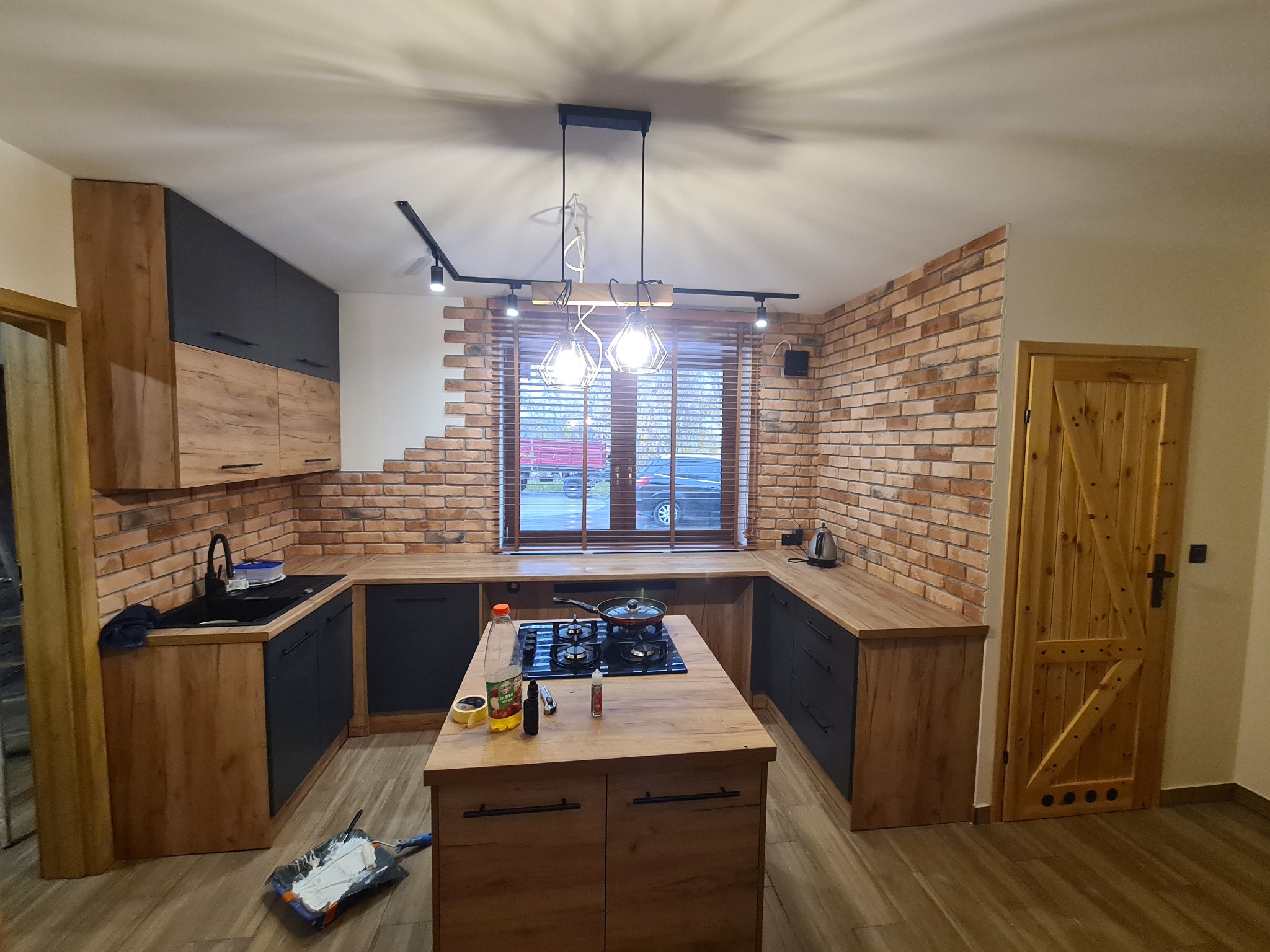 Kuchnia w stylu rustykalnym z ceglaną ścianą, drewnianymi blatami i czarnymi szafkami, wyspa kuchenna z płytą indukcyjną i stojąca na niej patelnia, widoczny wałek do malowania na podłodze.