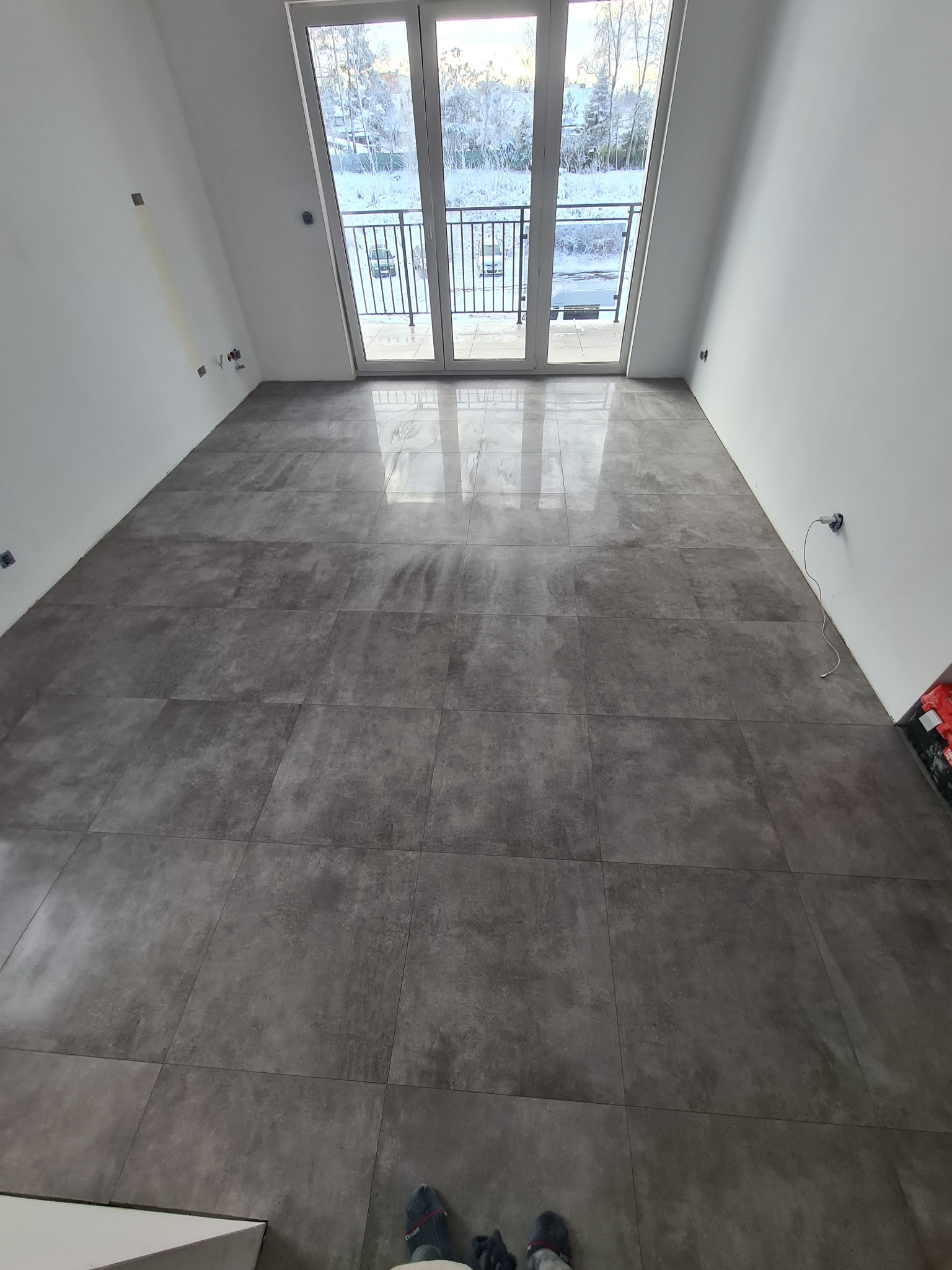 Ułożona, ciemnoszara, błyszcząca podłoga z płytek w nowym pomieszczeniu z dużym oknem balkonowym wychodzącym na zimowy krajobraz. Widoczne ślady po montażu na ścianach i fragment butów w dolnej...