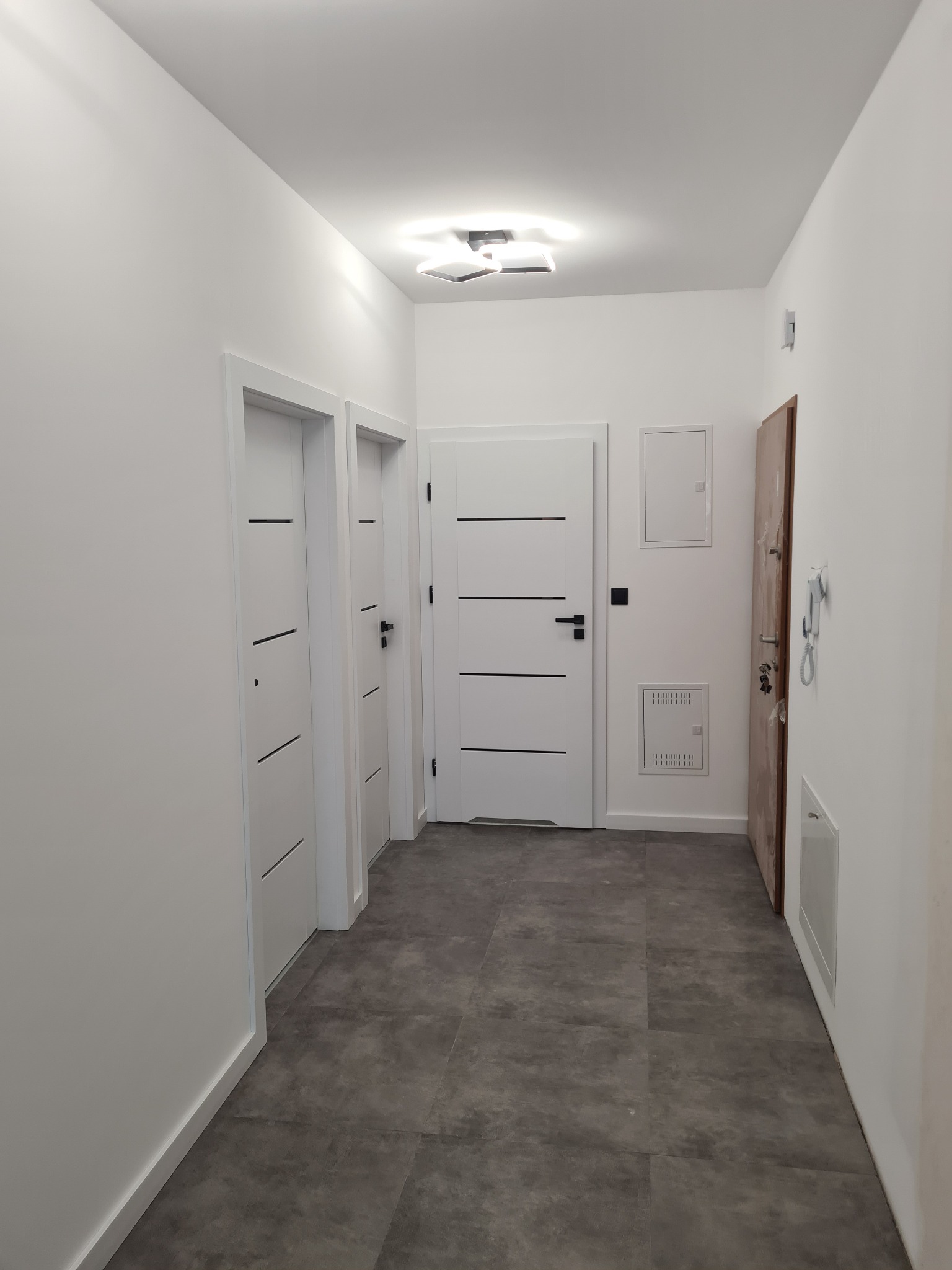 Jasny korytarz po remoncie z trzema białymi drzwiami z poziomymi frezami, szarą podłogą z płytek i nowoczesną lampą sufitową.