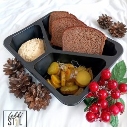 Lekki Styl Catering Dietetyczny - Catering dietetyczny: Czarna tacka z trzema przegródkami zawierająca hummus, chleb razowy i ogórki kiszone z kiełkami, udekorowana szyszkami i sztucznymi czerwonymi owocami na białym tle.