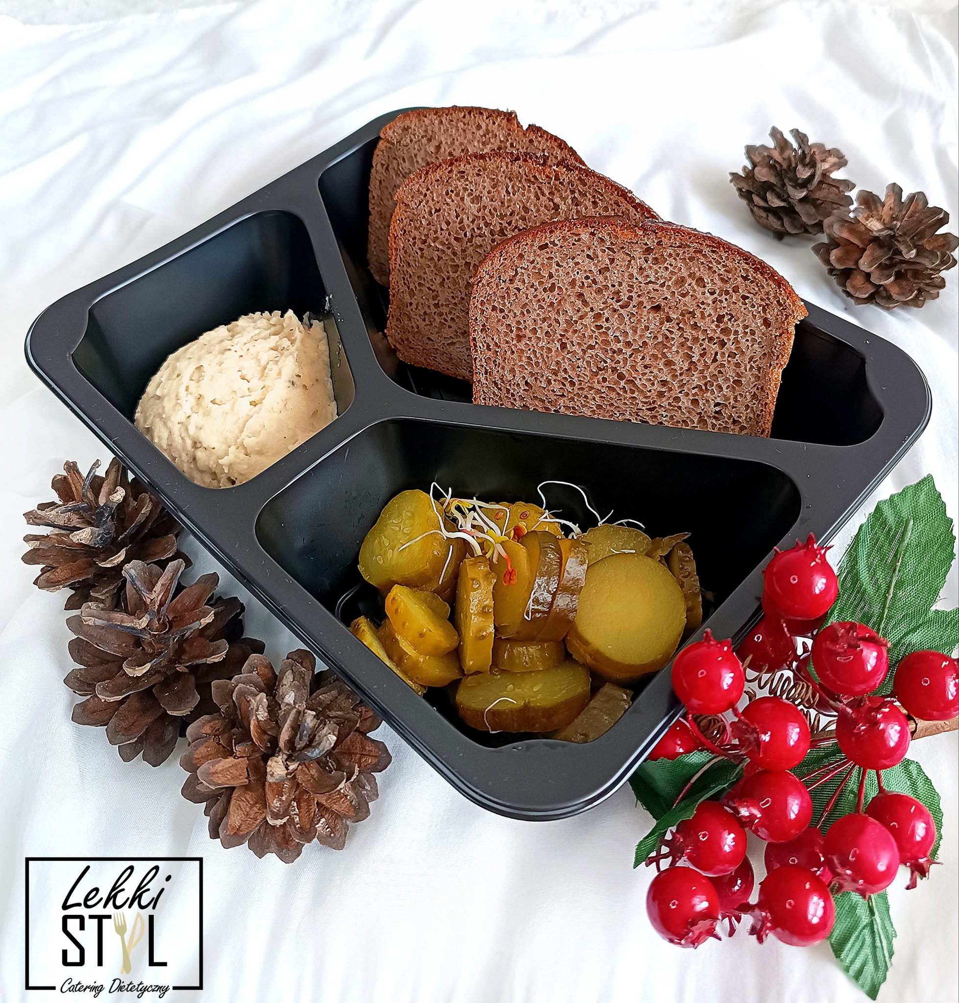 Catering dietetyczny: Czarna tacka z trzema przegródkami zawierająca hummus, chleb razowy i ogórki kiszone z kiełkami, udekorowana szyszkami i sztucznymi czerwonymi owocami na białym tle.
