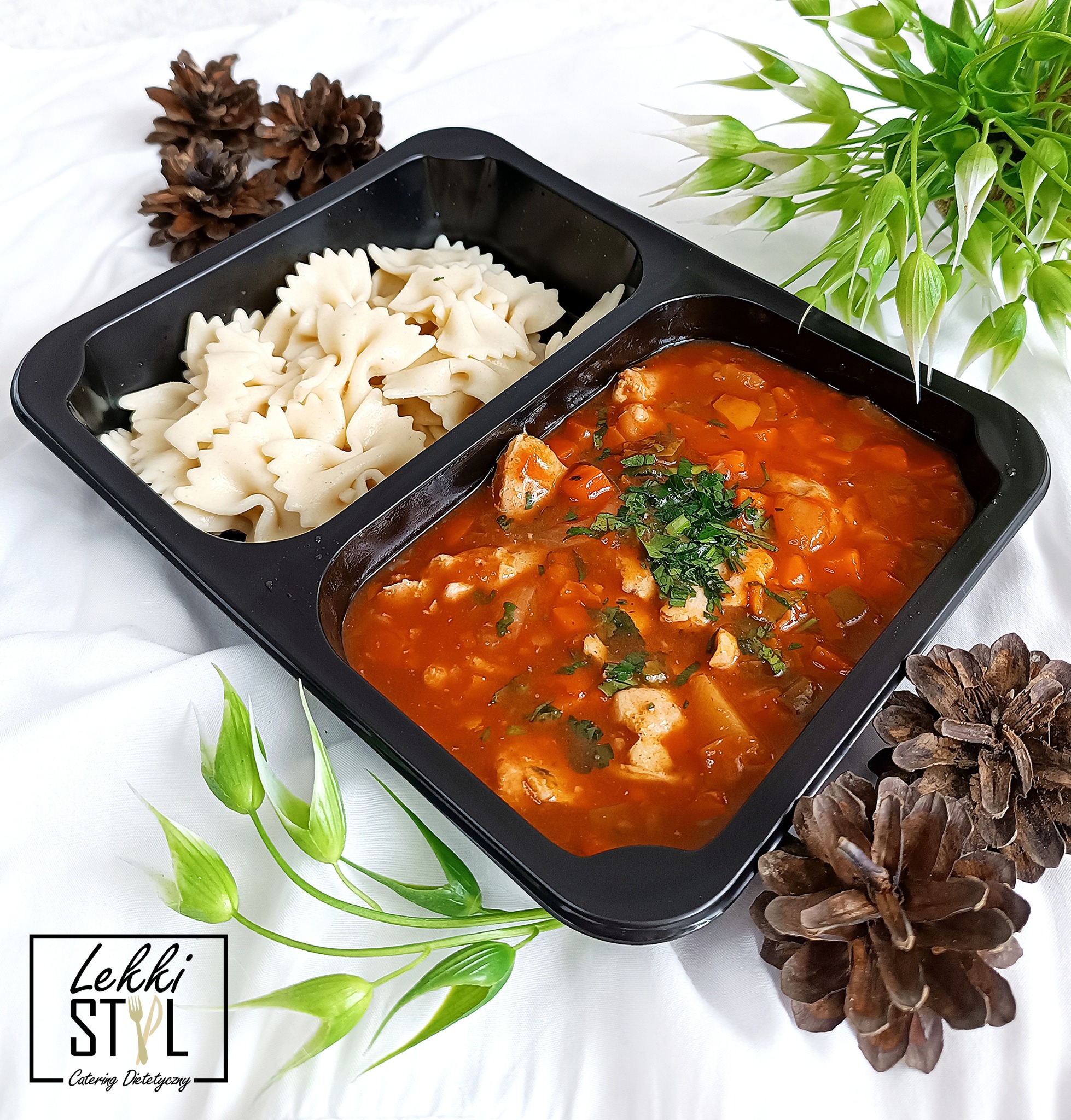 Gotowy posiłek w czarnym pudełku z dwoma przegródkami: makaron w kształcie kokardek i gulasz z kurczakiem i warzywami, udekorowany zieloną pietruszką, całość na białym tle z dekoracjami z szyszek...