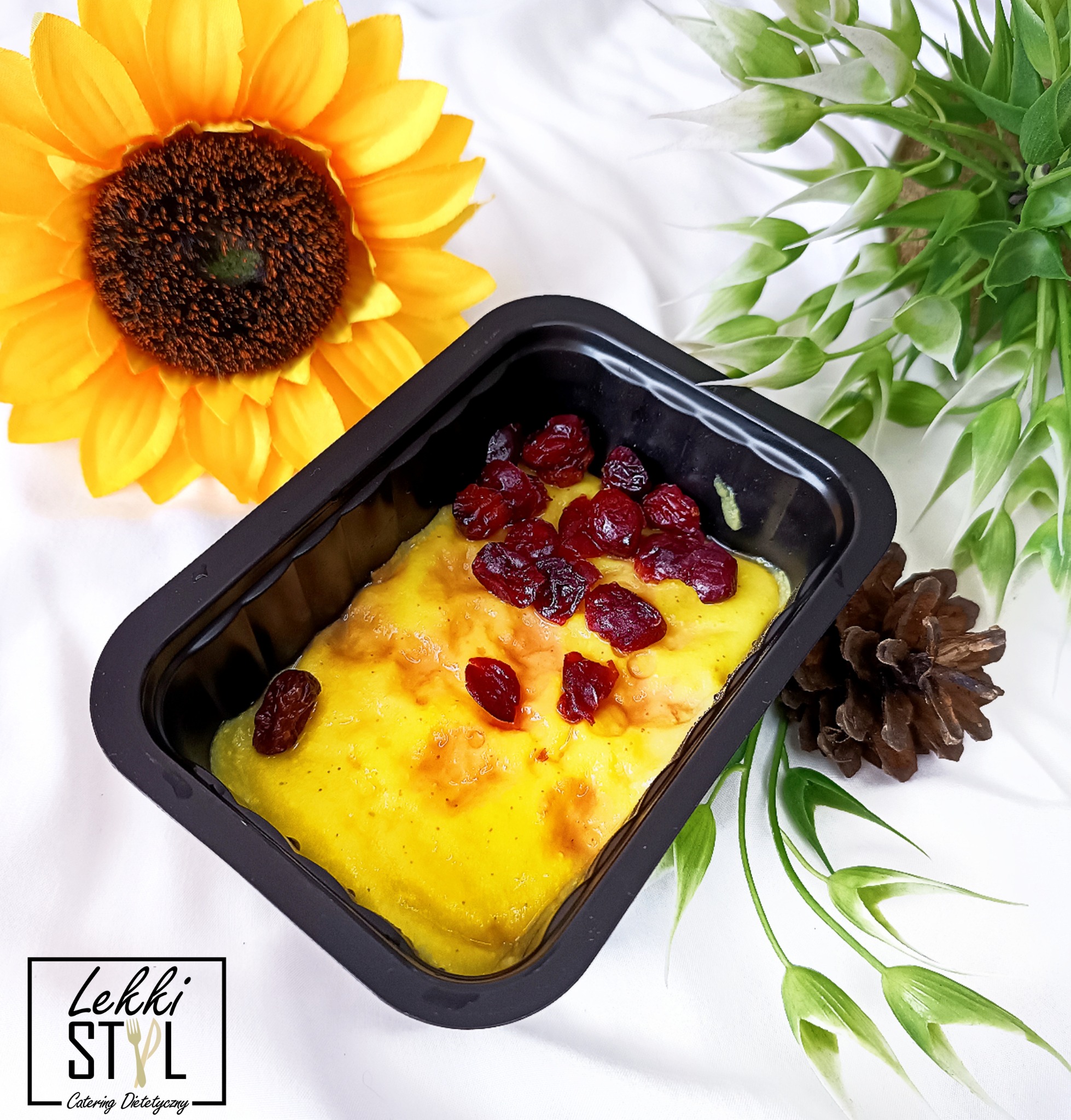 Danie z żółtym sosem i żurawiną w czarnym opakowaniu, otoczone dekoracjami: słonecznik, gałązki i szyszka. W lewym dolnym rogu logo firmy.