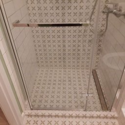 Kompleksowe Wykańczanie Łazienek - Szklana kabina prysznicowa z chromowanymi detalami, widok z góry, beżowe płytki z motywem kwiatowym na ścianach i podłodze, odpływ liniowy przy kabinie.