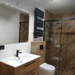 Kompleksowe Wykańczanie Łazienek - Nowoczesna łazienka z drewnopodobnymi płytkami na ścianach i podłodze, czarna armatura, umywalka z drewnianą szafką, kabina prysznicowa z czarnymi ramami, wisząca toaleta i czarny grzejnik...