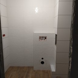 Kompleksowe Wykańczanie Łazienek - Wnętrze łazienki w trakcie remontu, widoczne białe płytki ścienne z delikatnym wzorem, drewnopodobne płytki podłogowe, przygotowane otwory na instalację sanitarną, surowy fragment ściany przy wejściu.