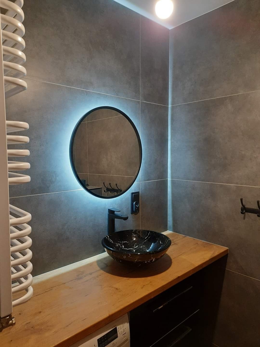 Nowoczesna łazienka z szarymi płytkami, okrągłym lustrem z podświetleniem LED, blatem z jasnego drewna i czarną, marmurkową umywalką nablatową.