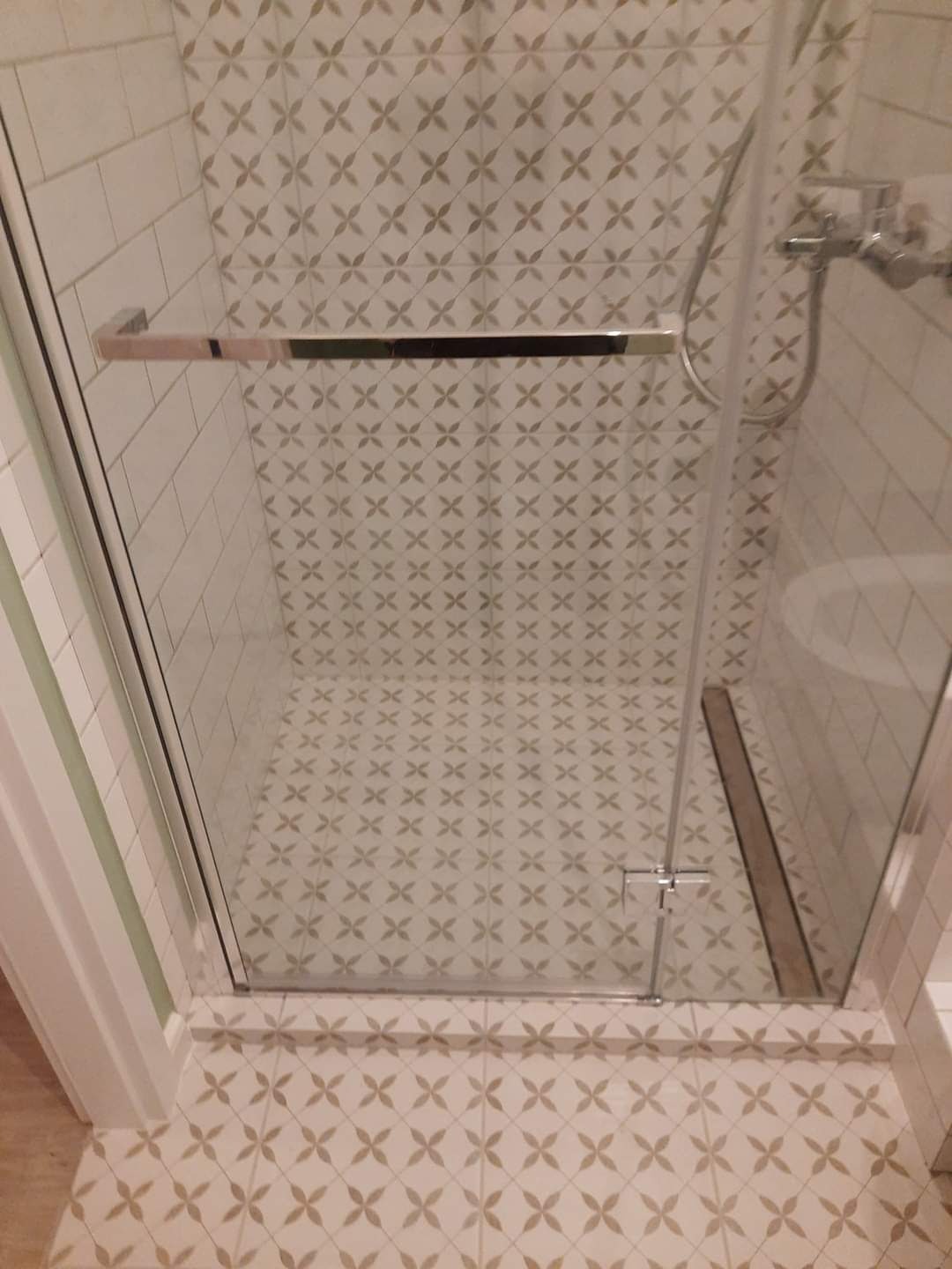 Szklana kabina prysznicowa z chromowanymi detalami, widok z góry, beżowe płytki z motywem kwiatowym na ścianach i podłodze, odpływ liniowy przy kabinie.