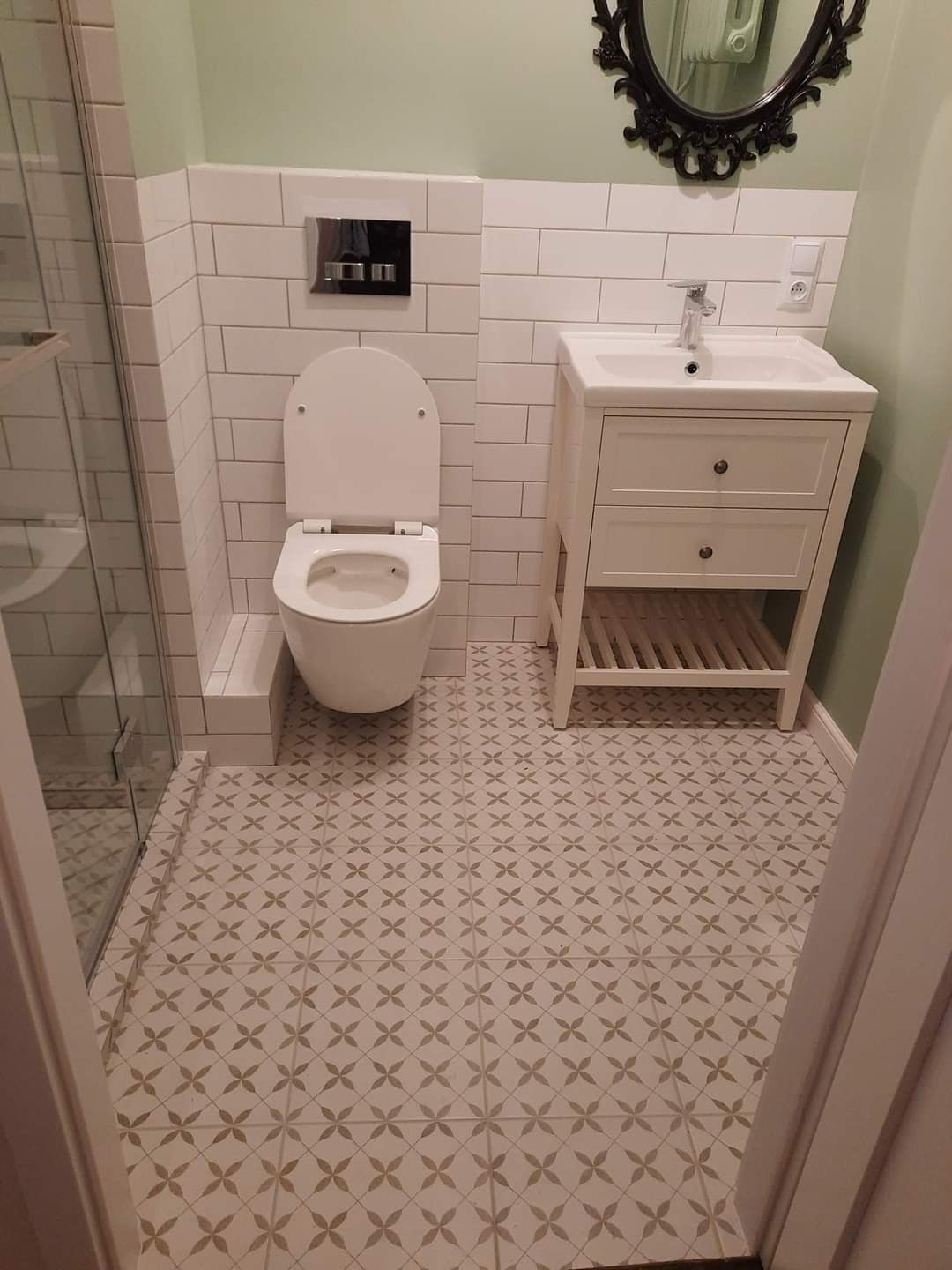 Wykończona łazienka z białymi płytkami imitującymi cegłę, toaletą wiszącą, białą szafką pod umywalkę z dwoma szufladami i wzorzystą podłogą w odcieniach szarości.