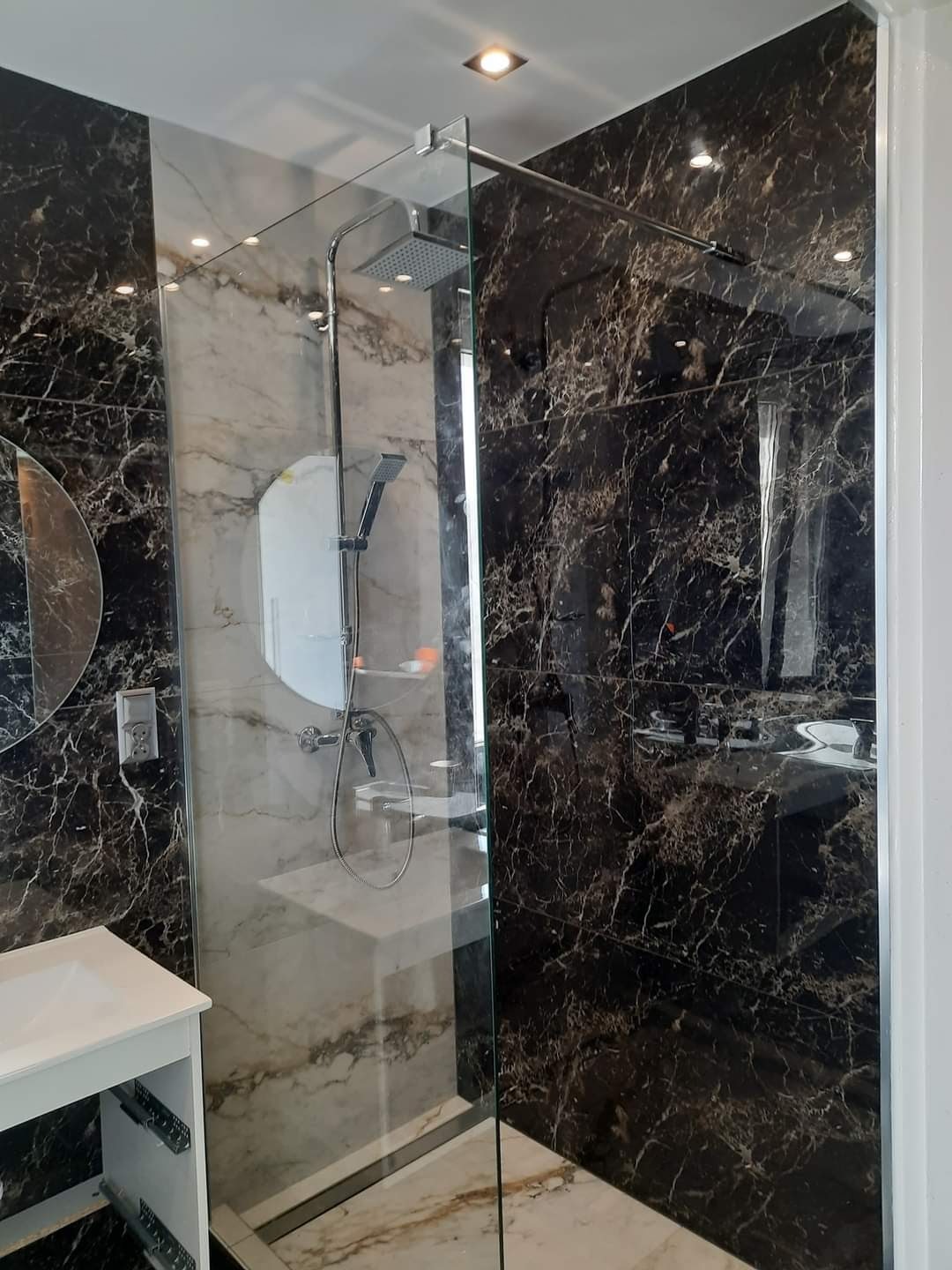 Nowoczesna kabina prysznicowa typu walk-in z czarnymi i białymi marmurkowymi płytkami, chromowaną armaturą i oświetleniem punktowym, widok z boku na umywalkę z szafką.