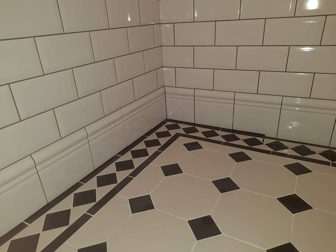 Wykończony narożnik łazienki z białymi, prostokątnymi płytkami ściennymi, dekoracyjnym cokołem ceramicznym i podłogą z heksagonalnych płytek w kolorze beżowym z czarnymi kwadratami.